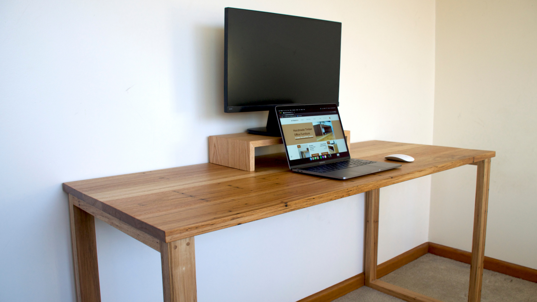 A wooden desk with a wall-mounted TV above it, a laptop, and a computer mouse on the desk in a room with beige walls and carpeted floor.
