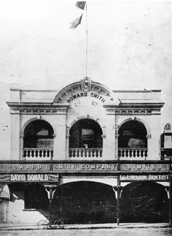 Old black and white building with sign 'Howard Smith Co Ltd', featuring three arched windows with decorative railings on the second floor and a small balcony. Multiple business signs on the ground floor include 'David Donald', 'G. L. Nelson Dentist', and 'Howard Smith Company Limited'.