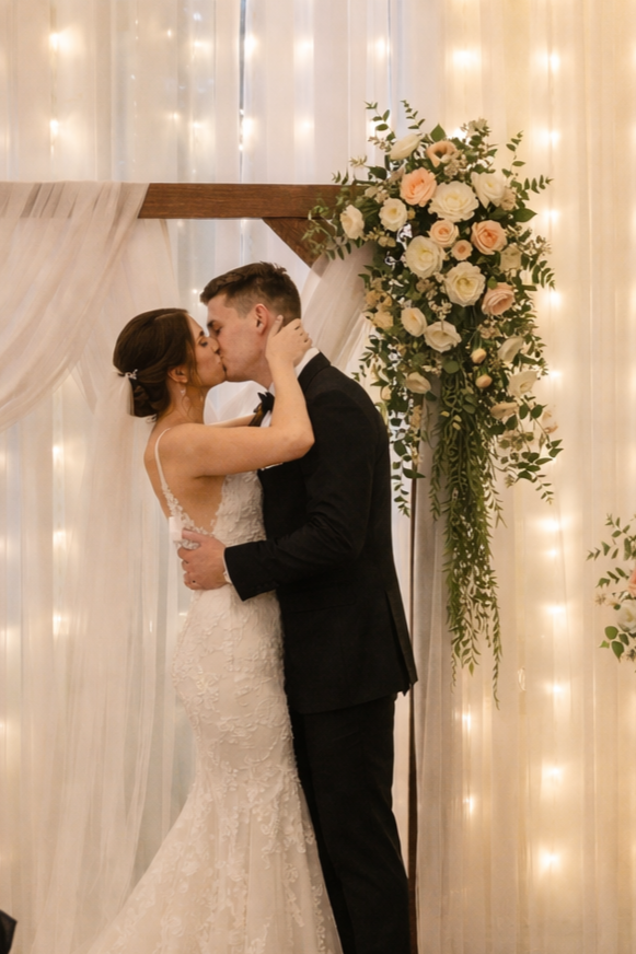 A bride and groom kissing at their wedding ceremony, standing in front of a floral arrangement with white and blush roses and greenery, adorned with string lights and draped fabric.