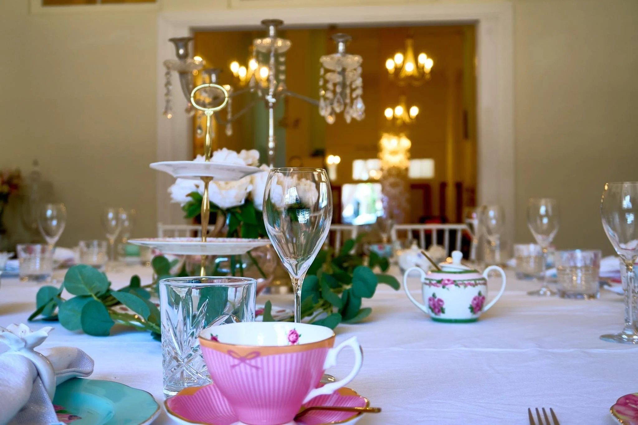 Elegant dining table set with glassware, floral centerpiece, teapot, and china cups, in a room with chandeliers and warm lighting.