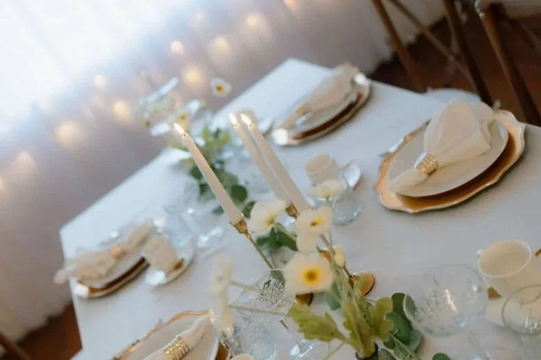 Decorated dining table with white candles, a floral centerpiece, and elegant place settings with folded napkins and plates.