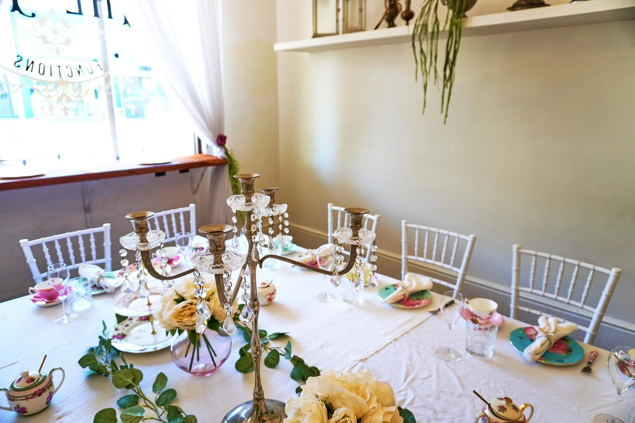 A dining table decorated for a celebration with a silver candelabrum, floral centerpiece, and pastel-colored tableware. The room has a window with sheer white curtains and a shelf with plants and decor above the wall.
