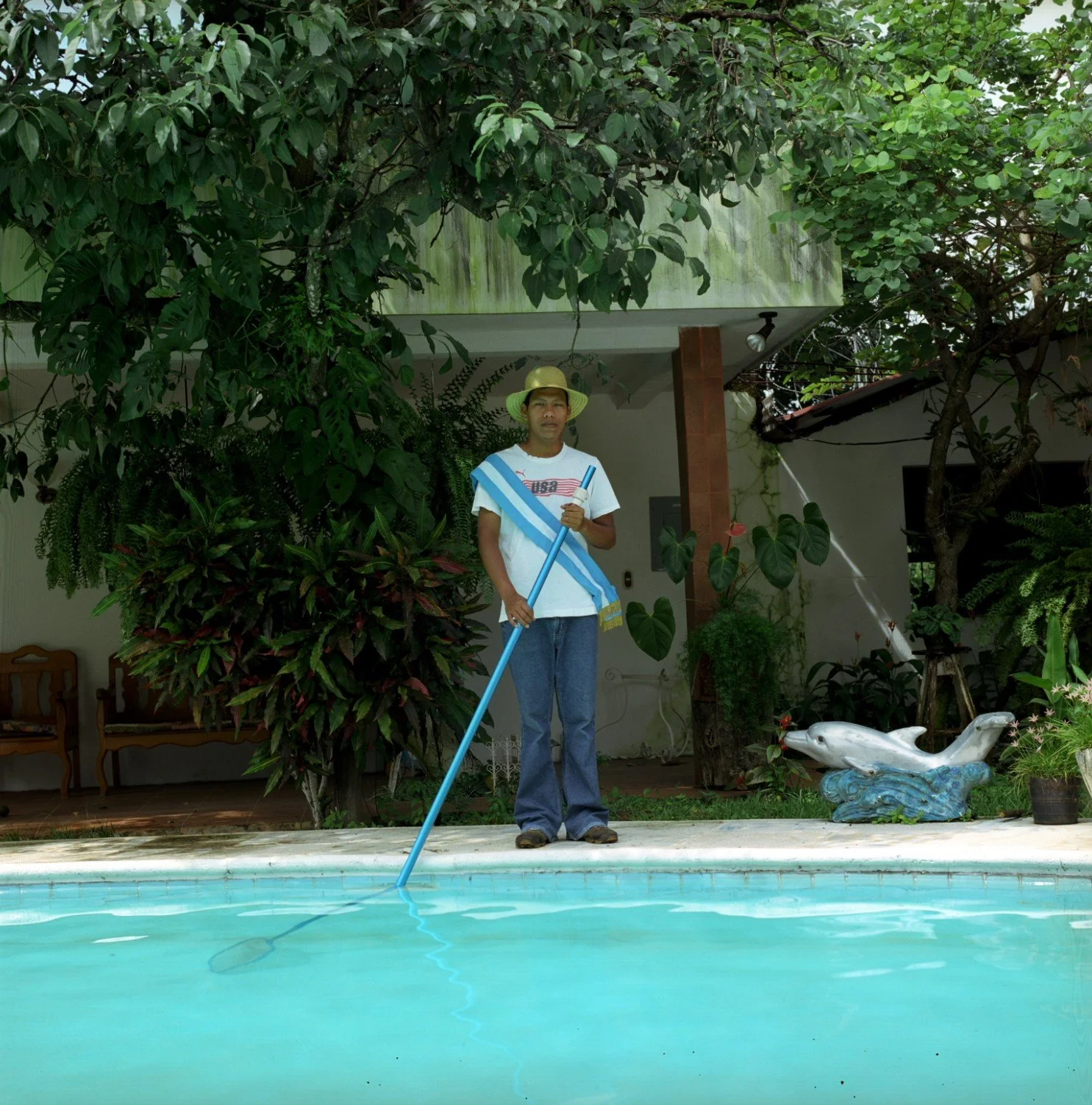 Joven con sombrero y camiseta blanca limpiando la piscina con una pértiga, en un entorno con plantas y una decoración de pez en la orilla.