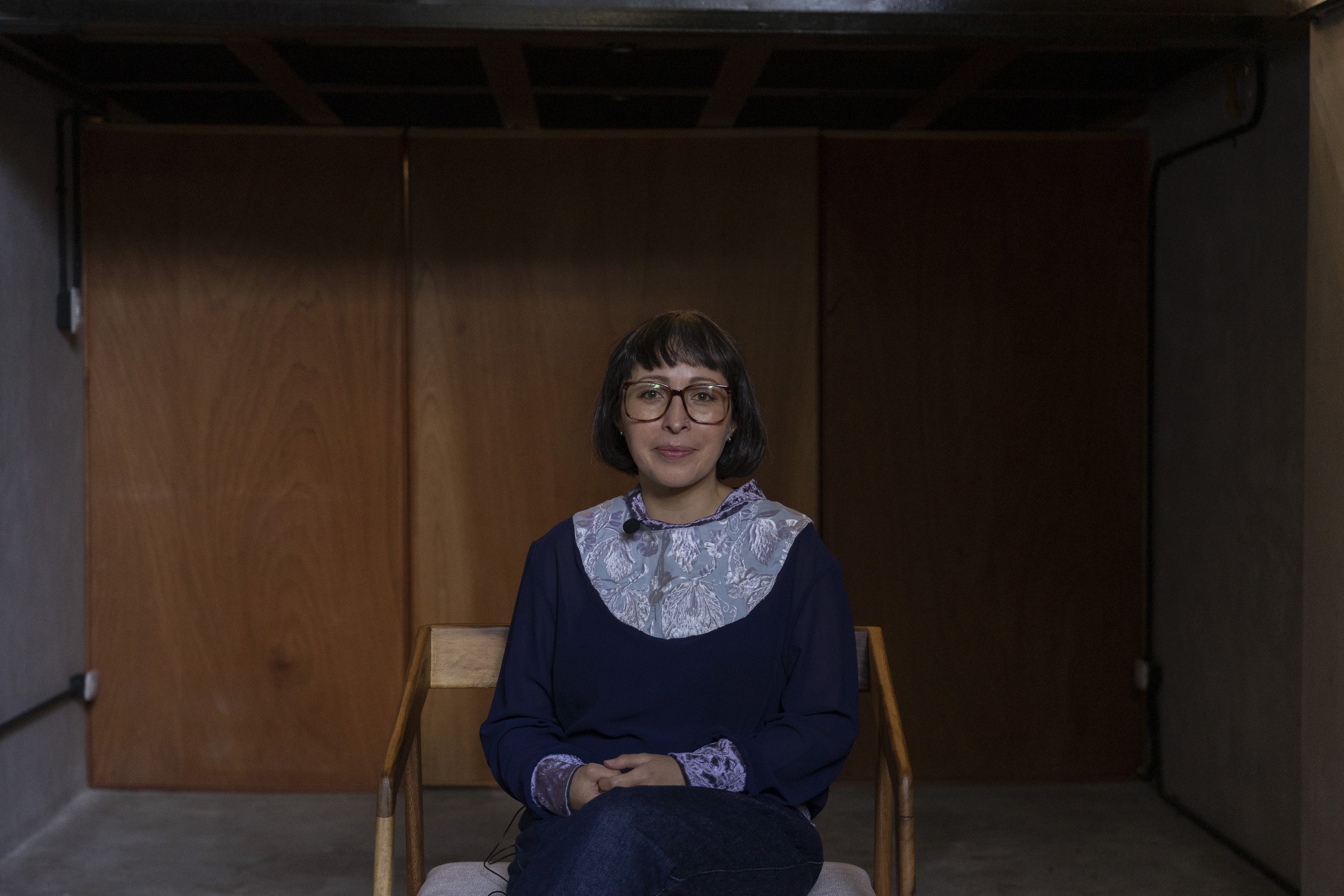 Mujer con gafas y vestido azul sentado en una silla en una habitación con paredes de madera