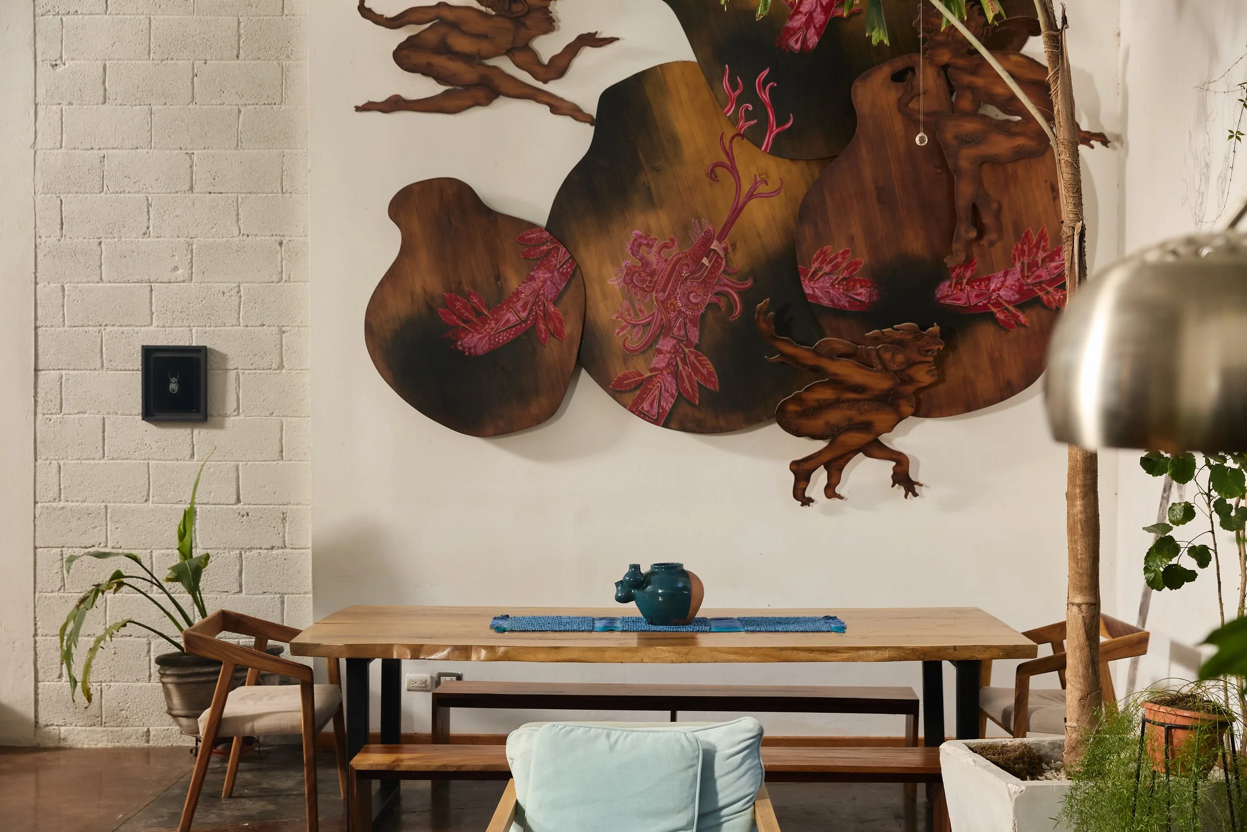 Decoración moderna en un comedor, con una pared blanca de fondo y una artística escultura de madera con detalles en color rosa y púrpura, acompañada de plantas y mobiliario de madera.