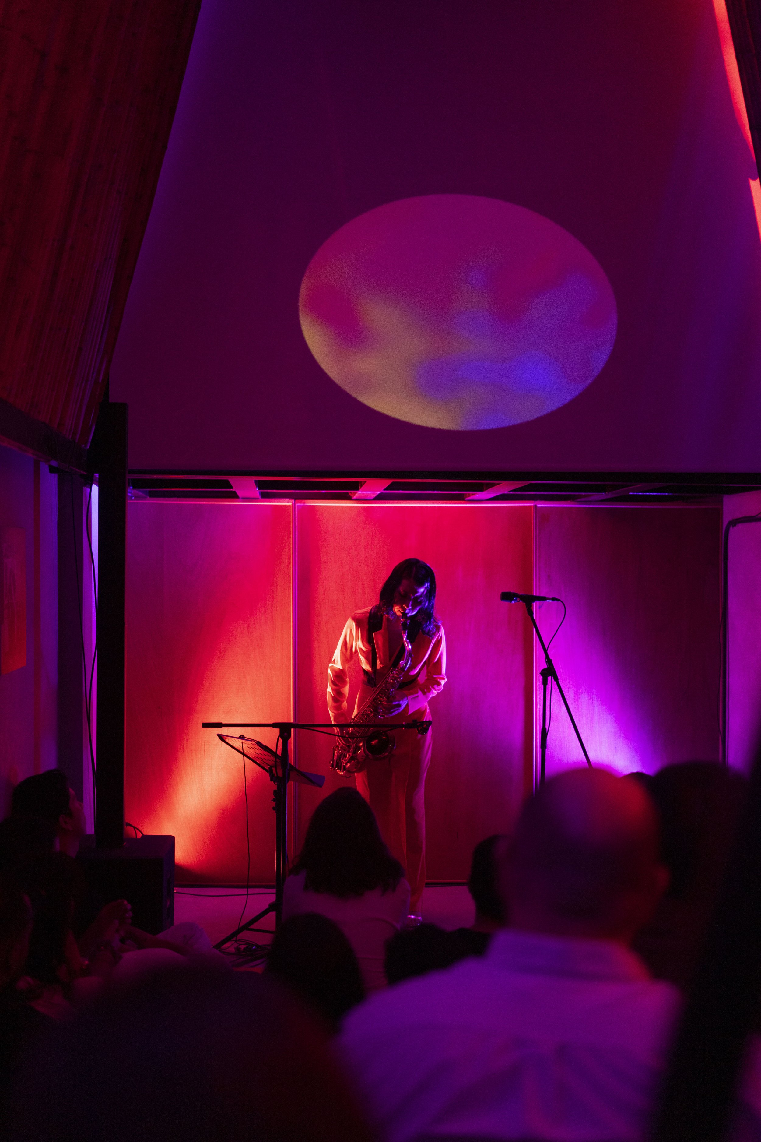 Un músico tocando el saxofón en un escenario con iluminación roja y púrpura, y una proyección de un círculo con efectos de color en la pared de fondo, mientras una audiencia observa.