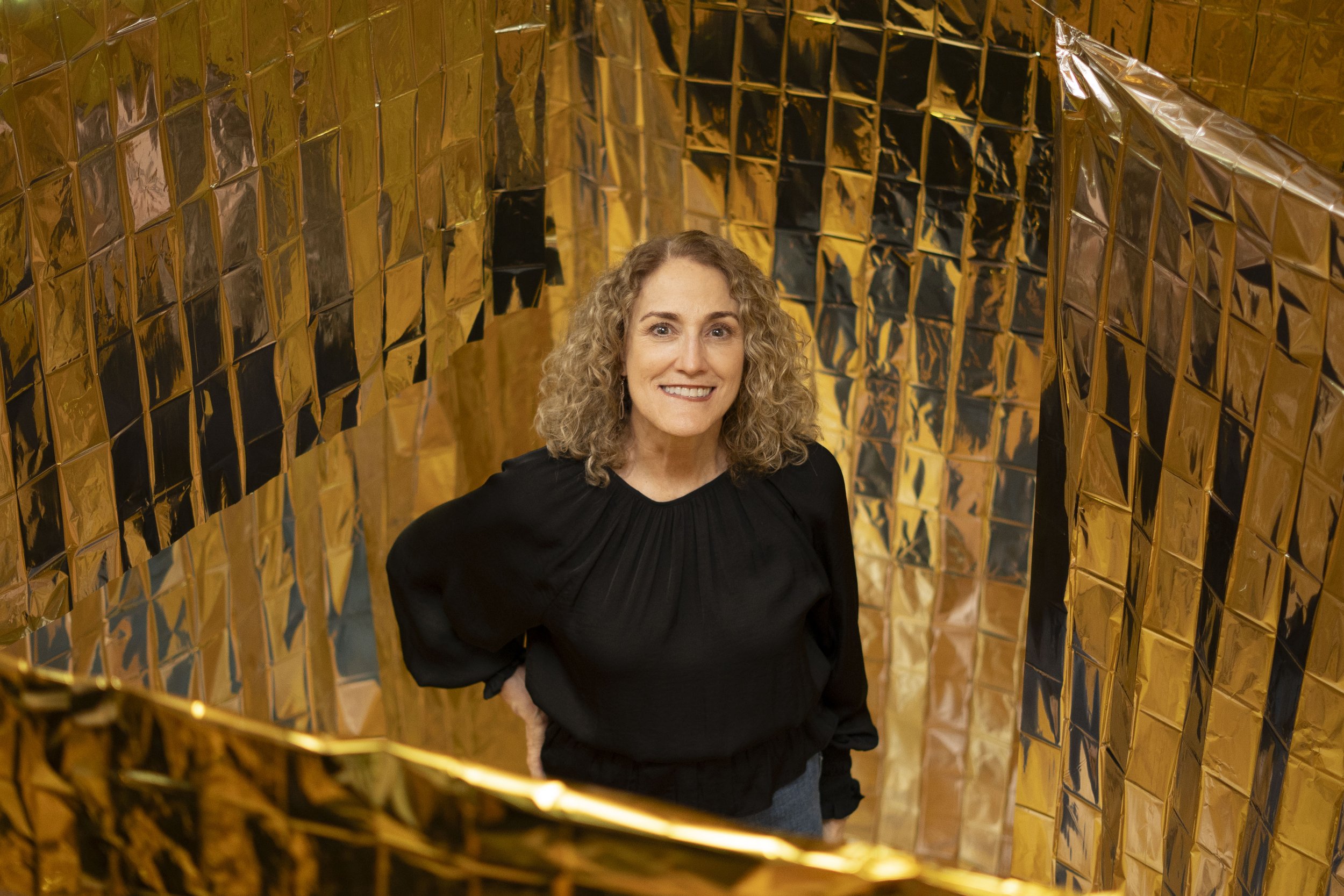Mujer de cabello rizado y rubio, sonriendo, de negro con fondo de paredes doradas reflectantes.