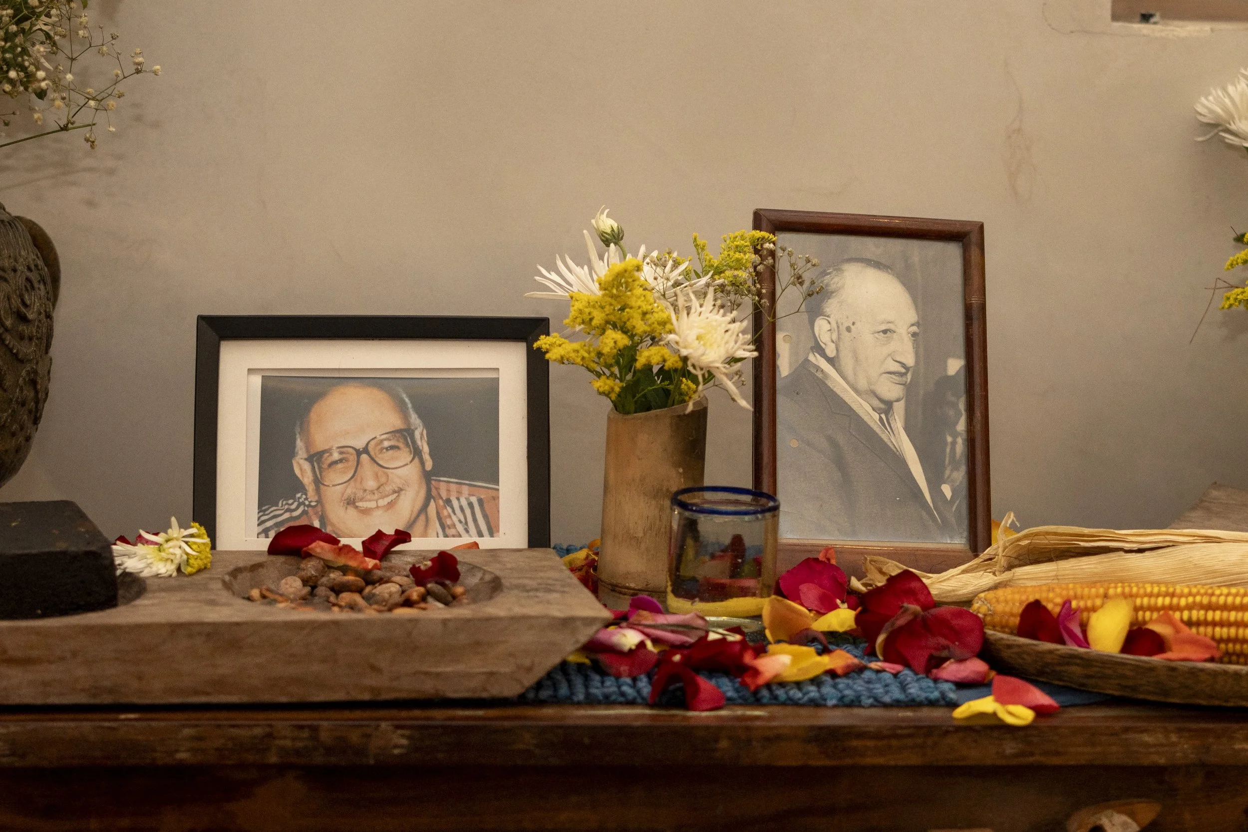 Dos fotografías enmarcadas en una mesa con una florero con flores, una vela, petalos y maíz cocido, en un altar dedicado a memoria de personas fallecidas.