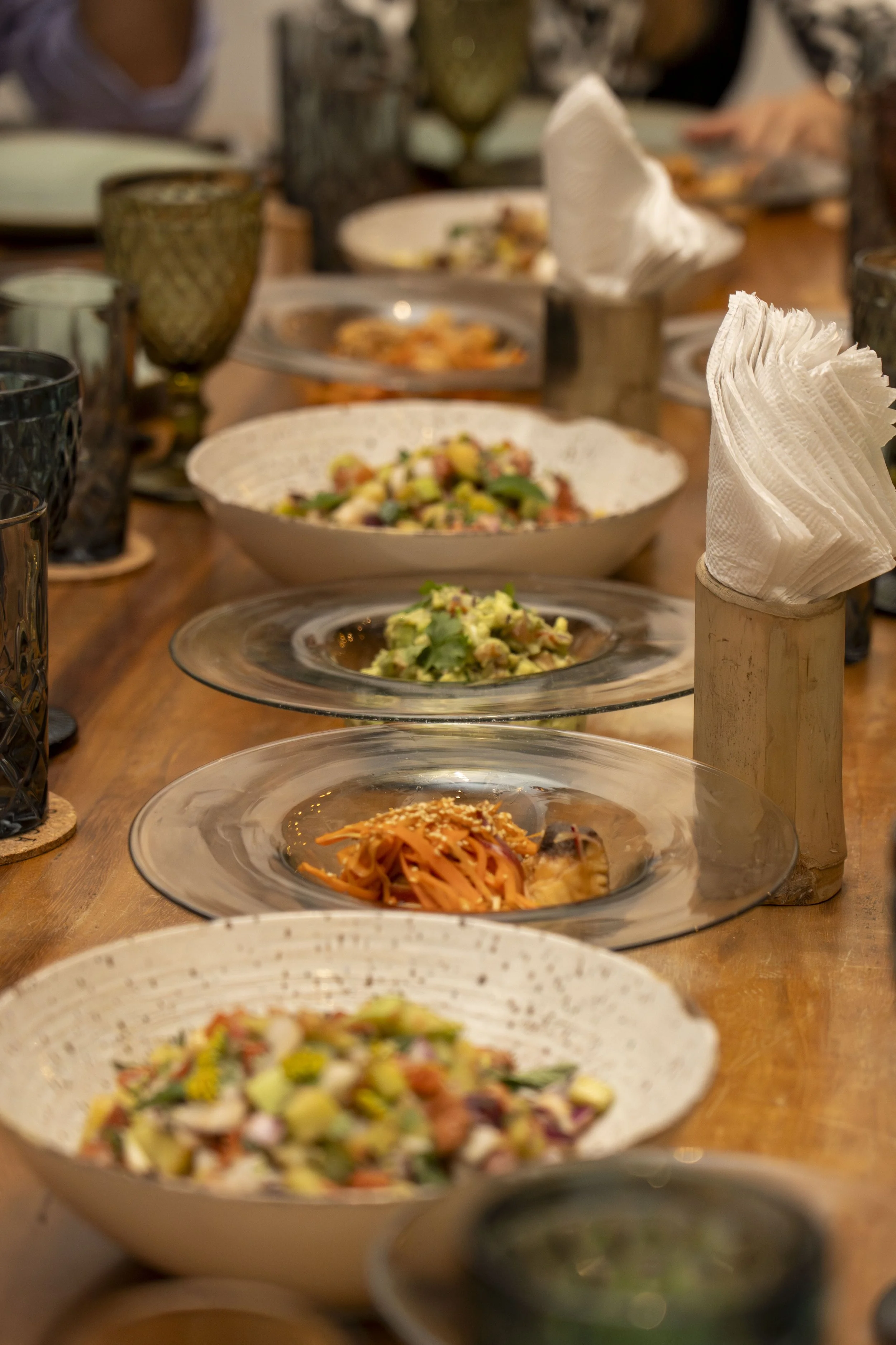 Una mesa con varios platos de ensaladas coloridas y pasta, junto con vasos de vidrio y paquetes de servilletas de papel.