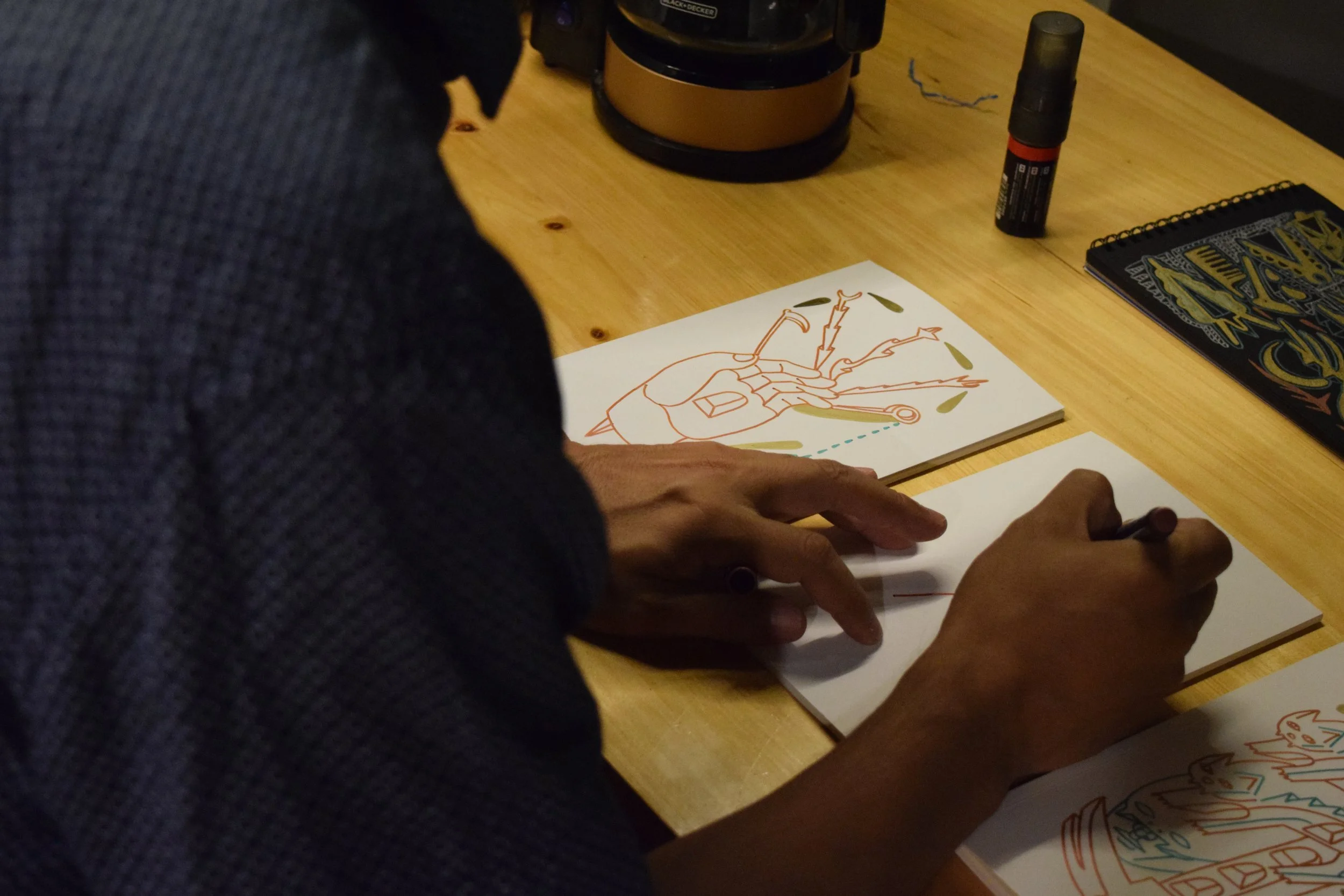 Persona dibujando un esquema de un robot con partes y conexiones en un cuaderno sobre una mesa de madera rodeada de material de dibujo y objetos