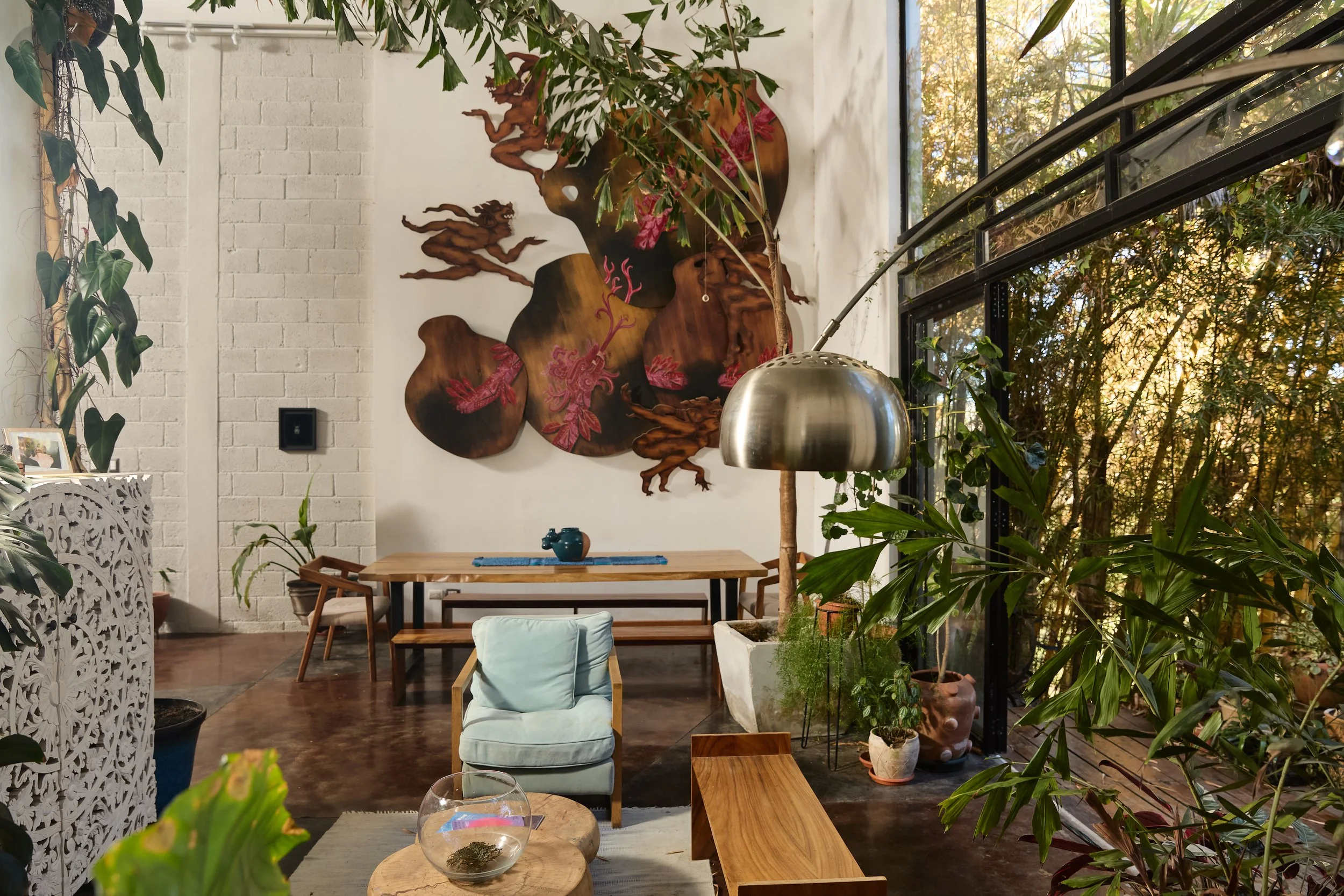 Interiores de un espacio con decoración moderna y plantas, con una pared decorada con arte en relieve y una mesa de madera con una tetera, en un ambiente iluminado por luz natural a través de grandes ventanas.