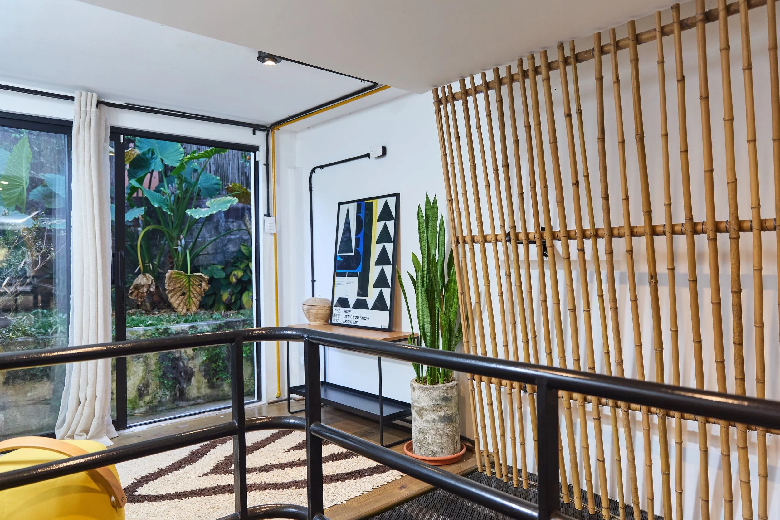 Interior de una sala con plantas, una pared de bambú decorativa, un marco con arte en blanco y negro, y una ventana con vista a un jardín con plantas grandes.