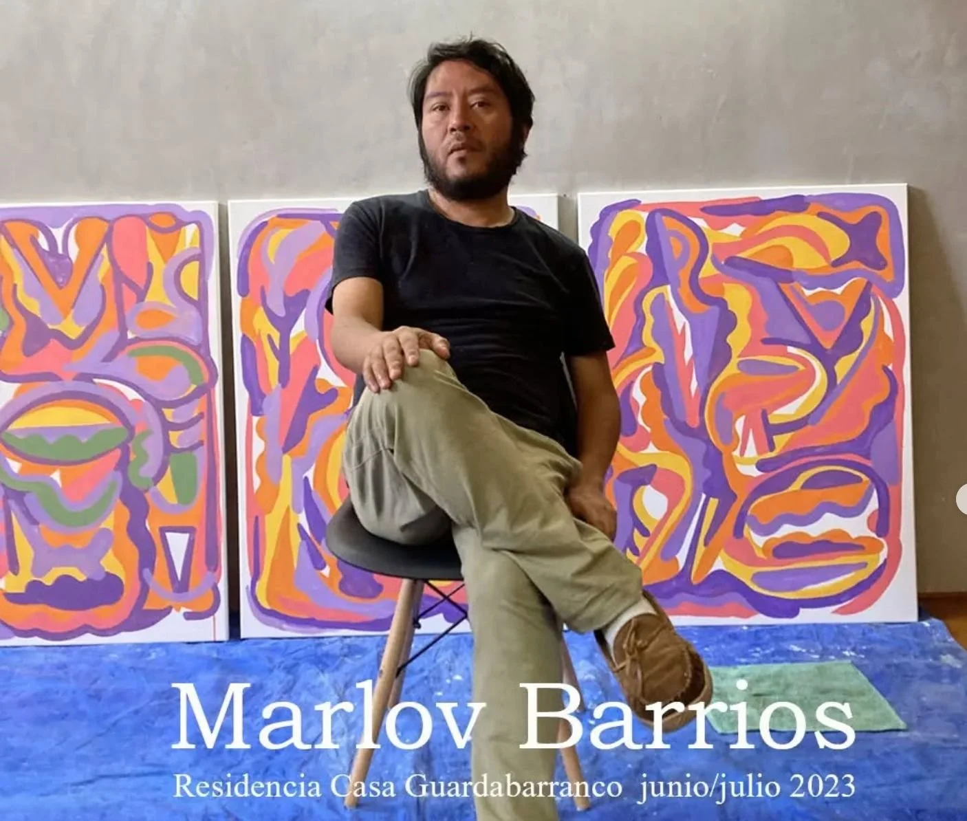 Un hombre sentado en una silla negro, usando una camiseta negra y pantalones beige, en una habitación con tres obras de arte abstracto en el fondo con colores vibrantes y formas orgánicas. Texto en la parte inferior que dice: "Marlov Barrios, Residencia Casa Guardabarranco, junio/julio 2023."