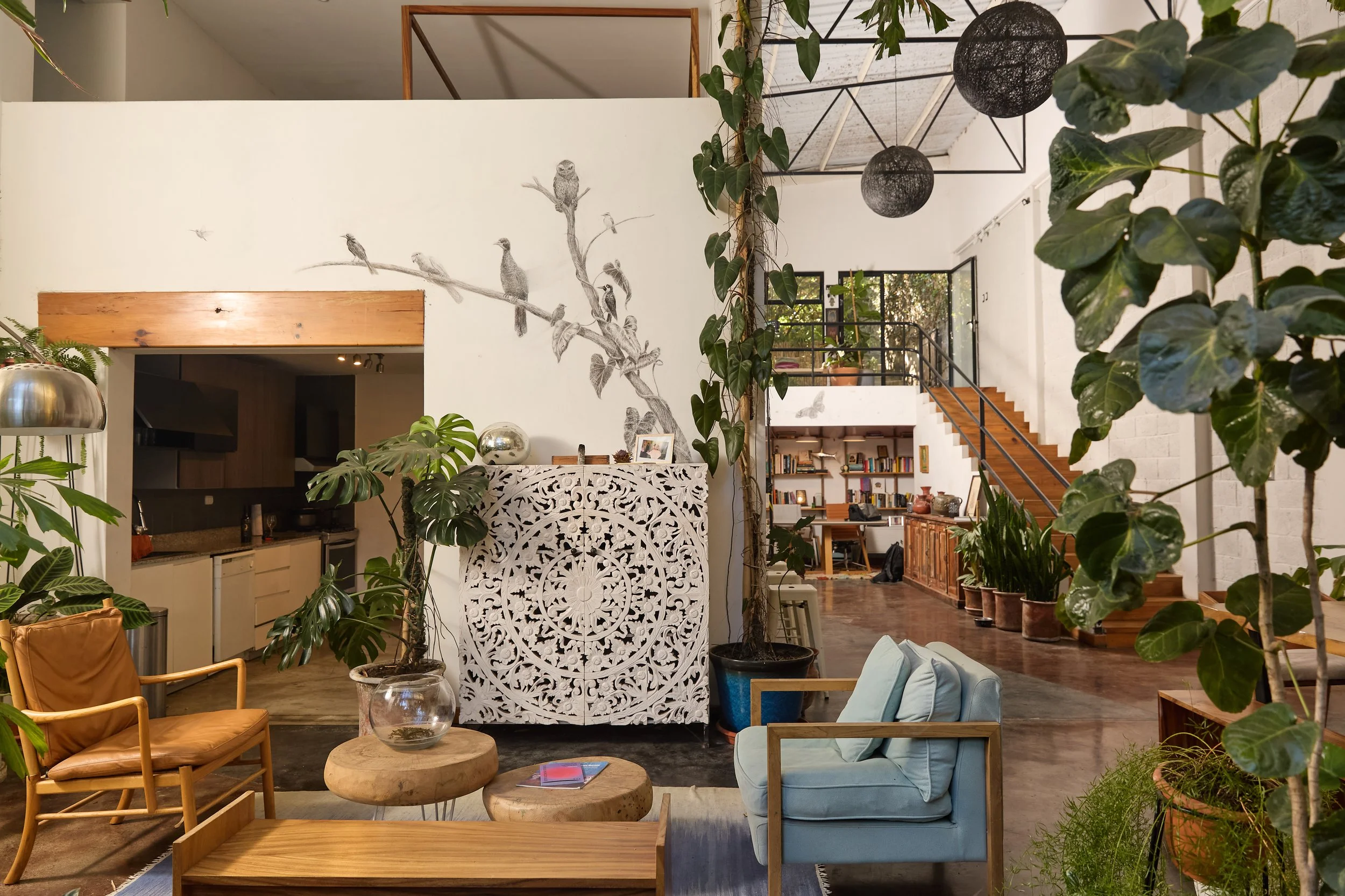 Interior de una sala de estar con muebles de madera, muchas plantas verdes, una pared decorada con un dibujo de aves y ramas, pasillo con estanterías llenas de libros, escaleras de madera y lámparas negras colgantes.