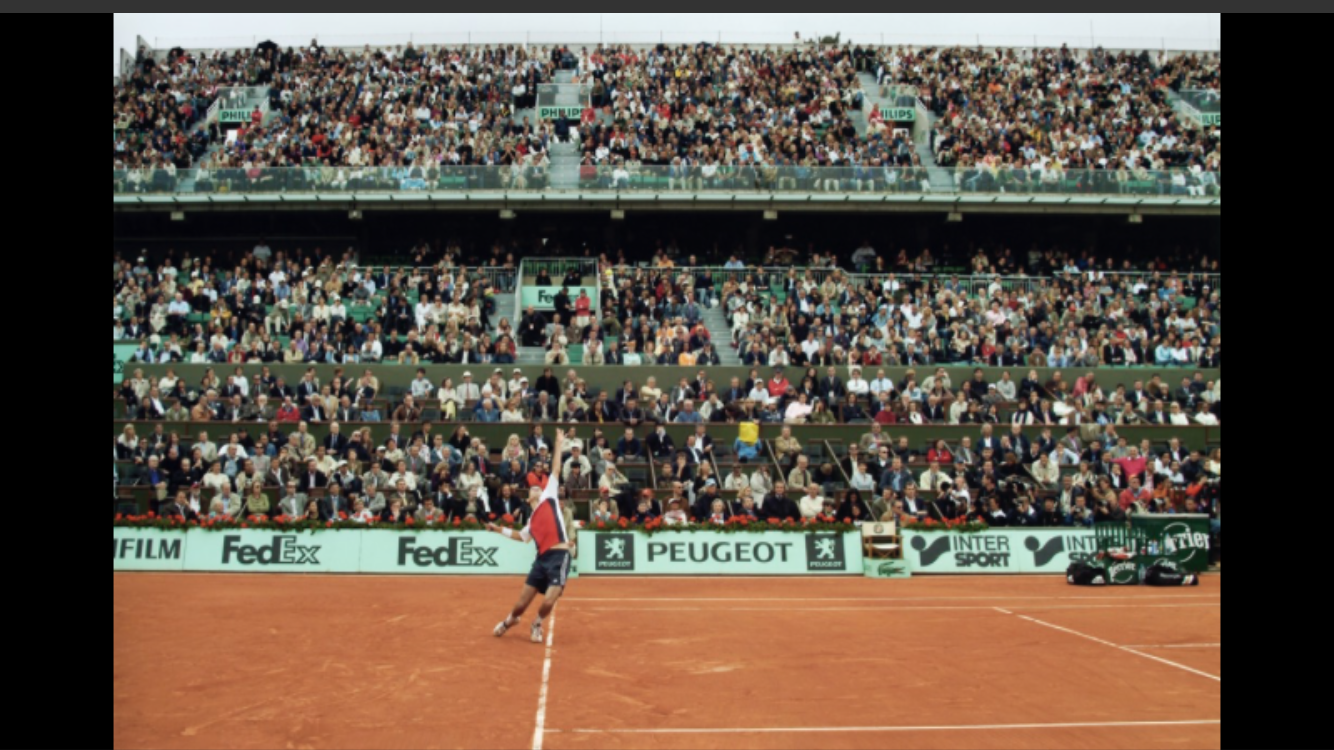 Jugador de tenis en la pista de arcilla, rodeado de espectadores en las gradas durante un partido.