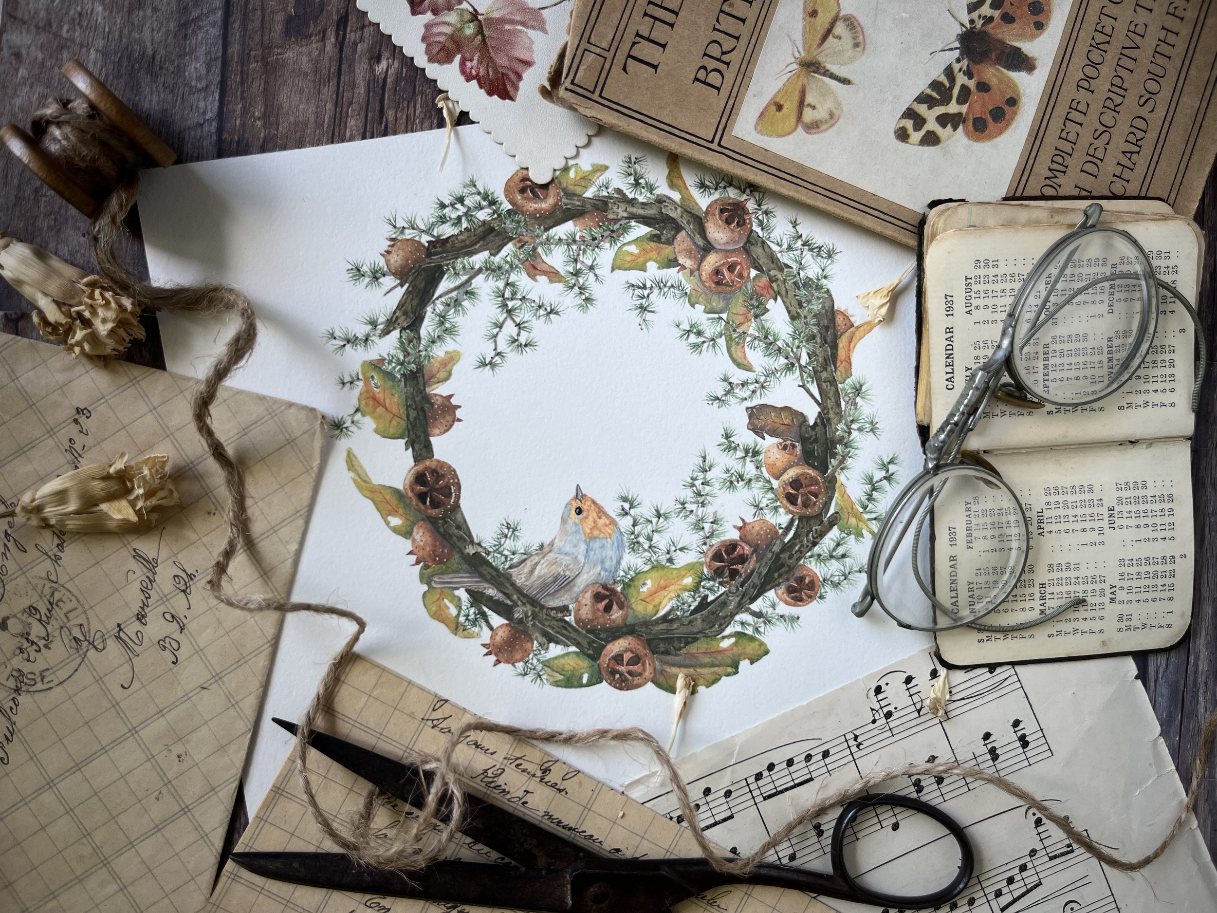 A collection of vintage items on a wooden surface, including a watercolor painting of a wreath with berries and leaves, a small bird, a pair of eyeglasses resting on an open calendar and a small book, handwritten notes, dried flowers, and a pair of b