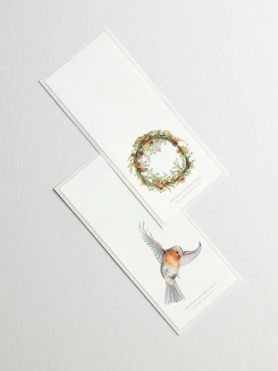 Two blank paper bookmarks, one with a floral wreath design and the other with a watercolor illustration of a bird in flight.
