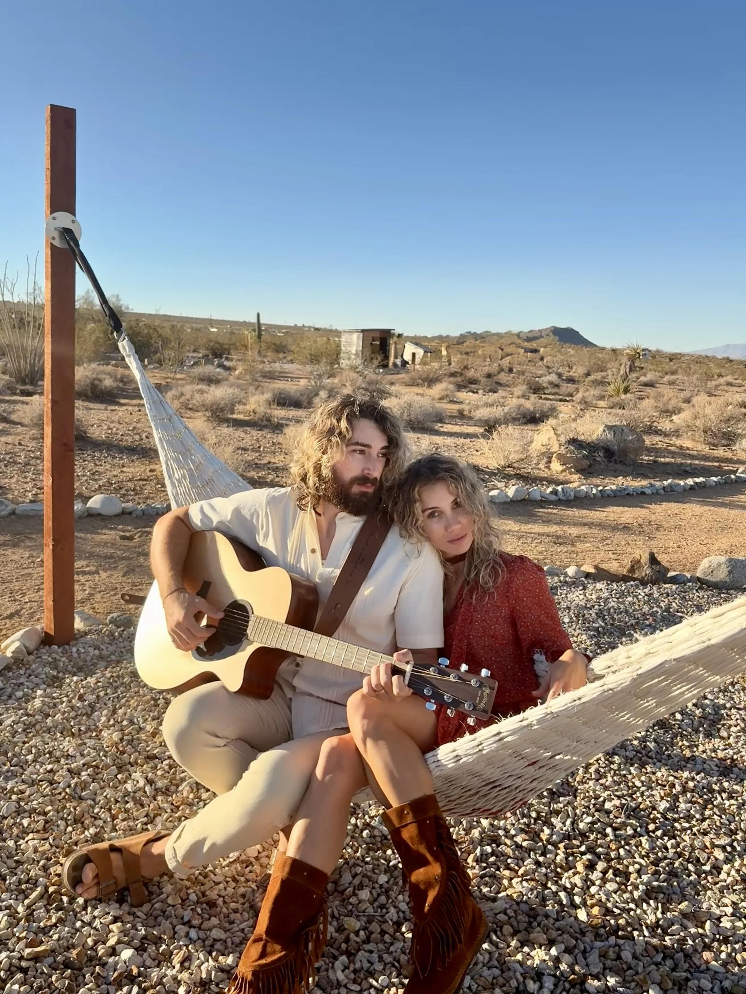 Bel Mercy promotional photo in the California desert featuring the husband-and-wife folk-rock duo with an acoustic guitar.
