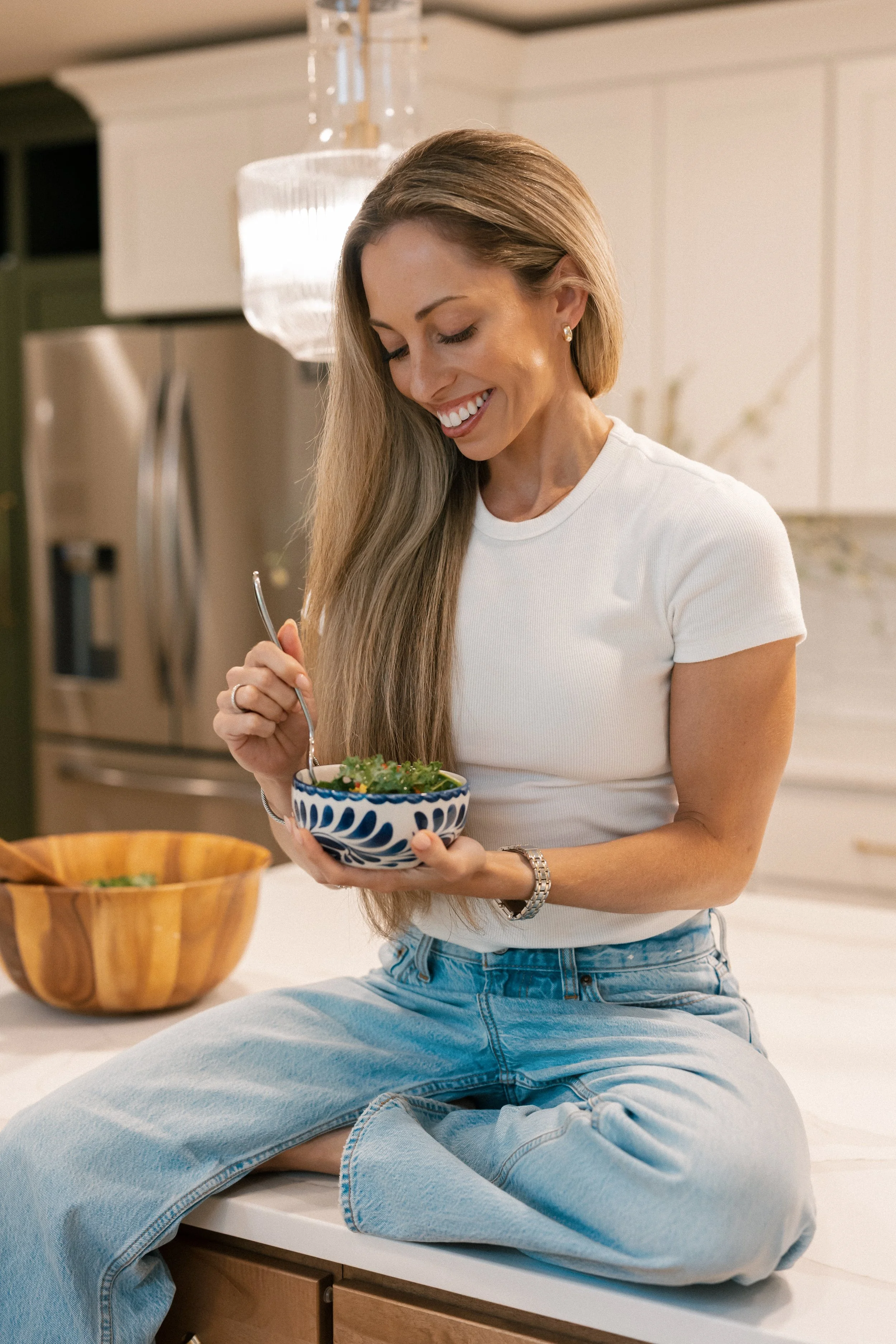 Lindsey with long blonde hair sitting on a kitchen counter, smiling while holding a bowl of salad.