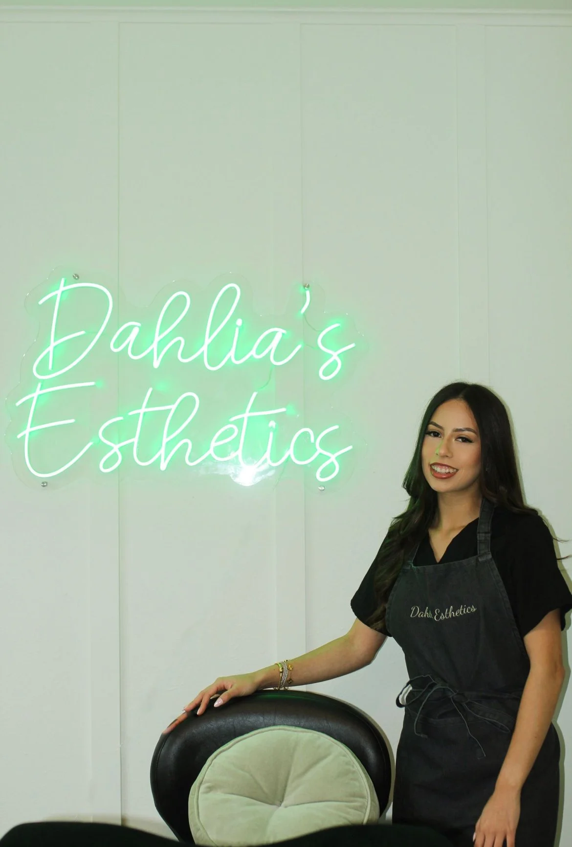 A woman in a black apron standing beside a black and beige chair in front of a white wall with a neon sign that reads 'Dahlia's Esthetics'.