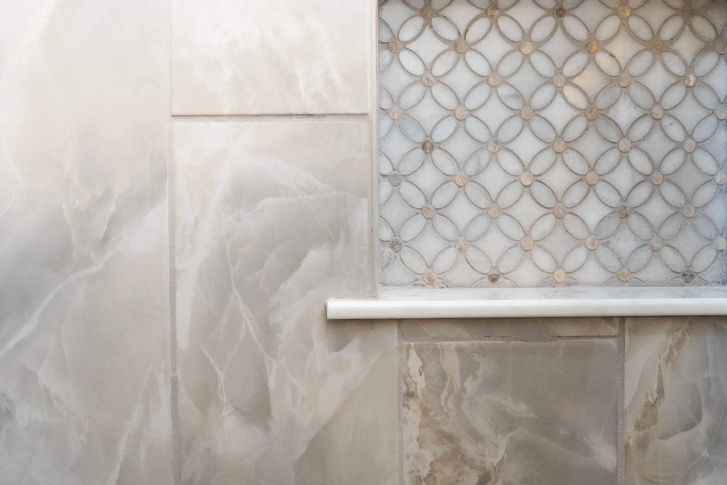 Close-up view of a tiled wall with two different styles of tiles: large beige marble tiles on the left and smaller decorative patterned tiles on the right, with a white ledge at the bottom.