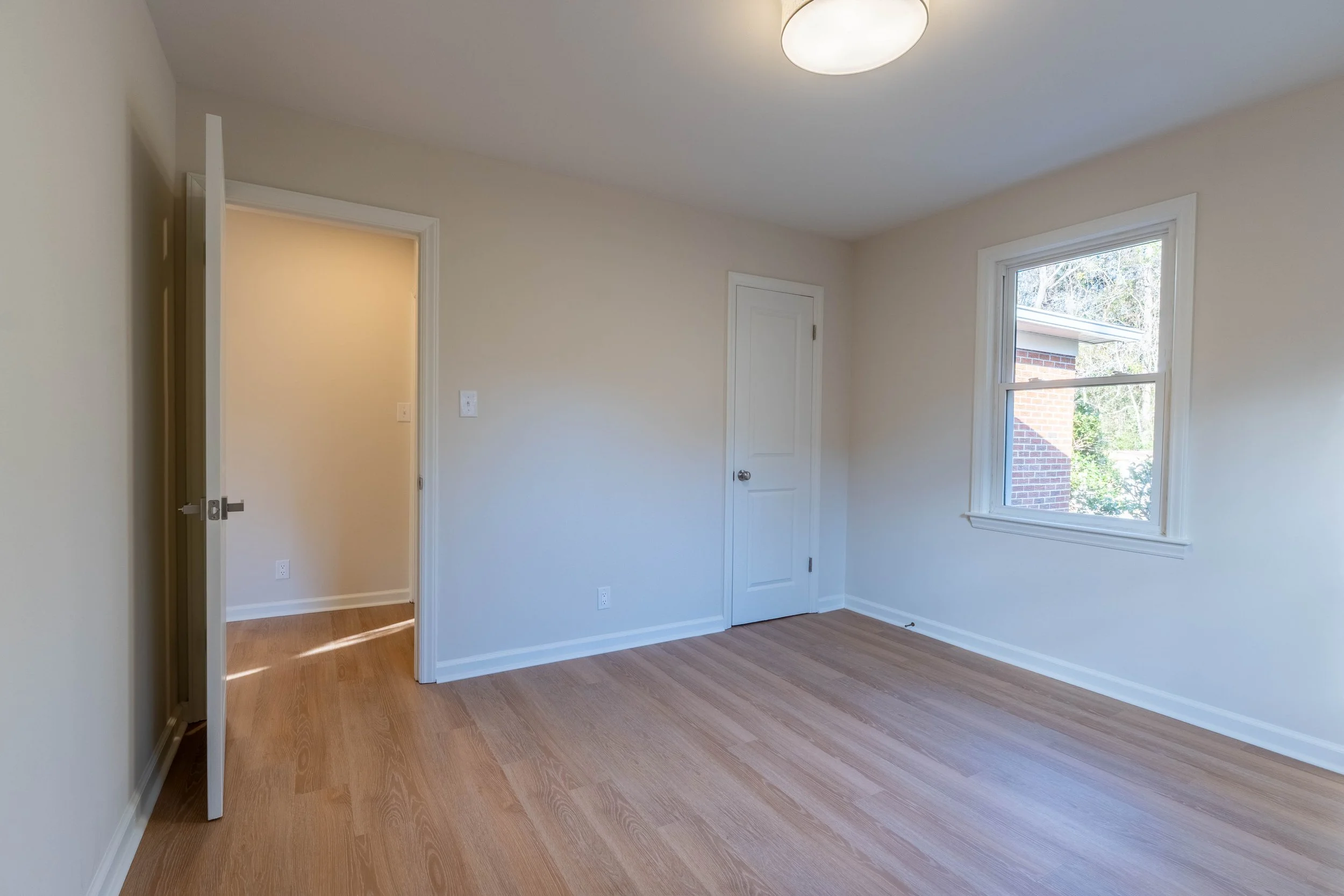 Empty room with hardwood floors, white walls, a window with a view of trees and brick building, a ceiling light, and a door.