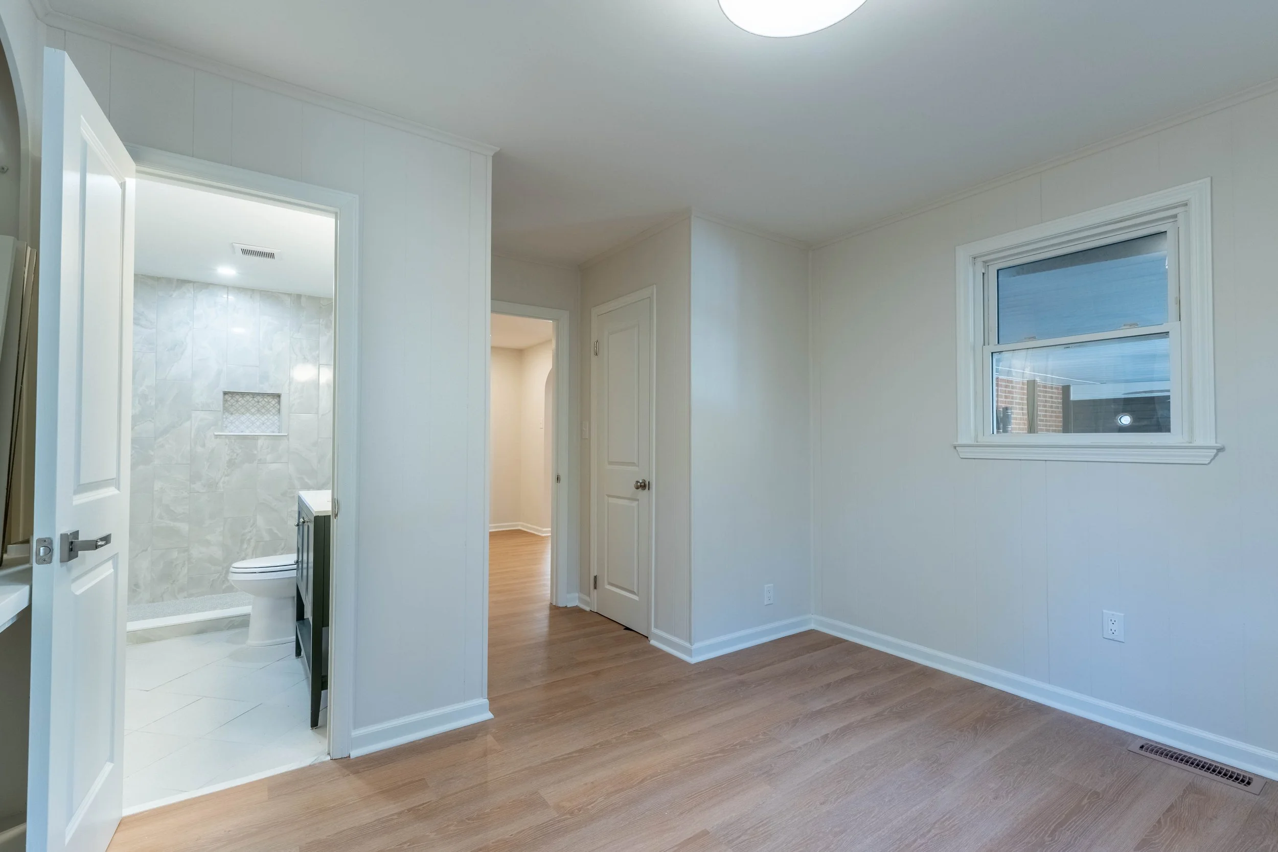 Empty room with white walls, wooden flooring, and a window showing a brick building outside. Doorways lead to a bathroom with a toilet and tiled walls.