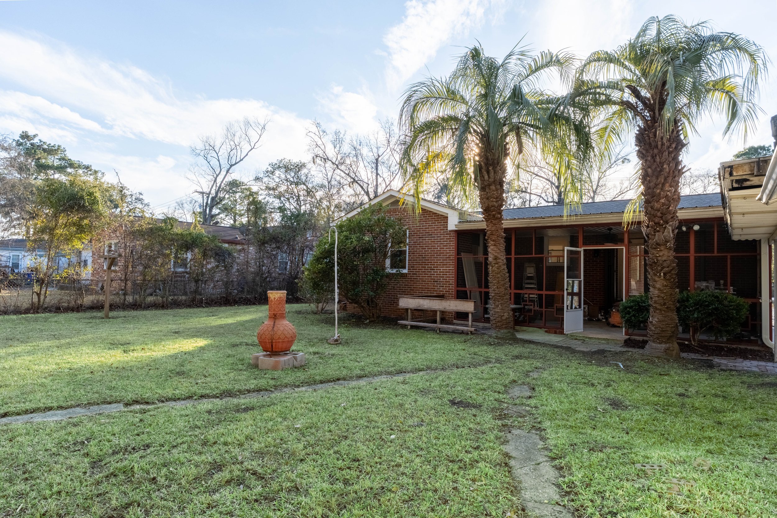 Backyard scene with green grass, a large clay pot, a brick house with a screened porch, and two tall palm trees with sunlight shining through.