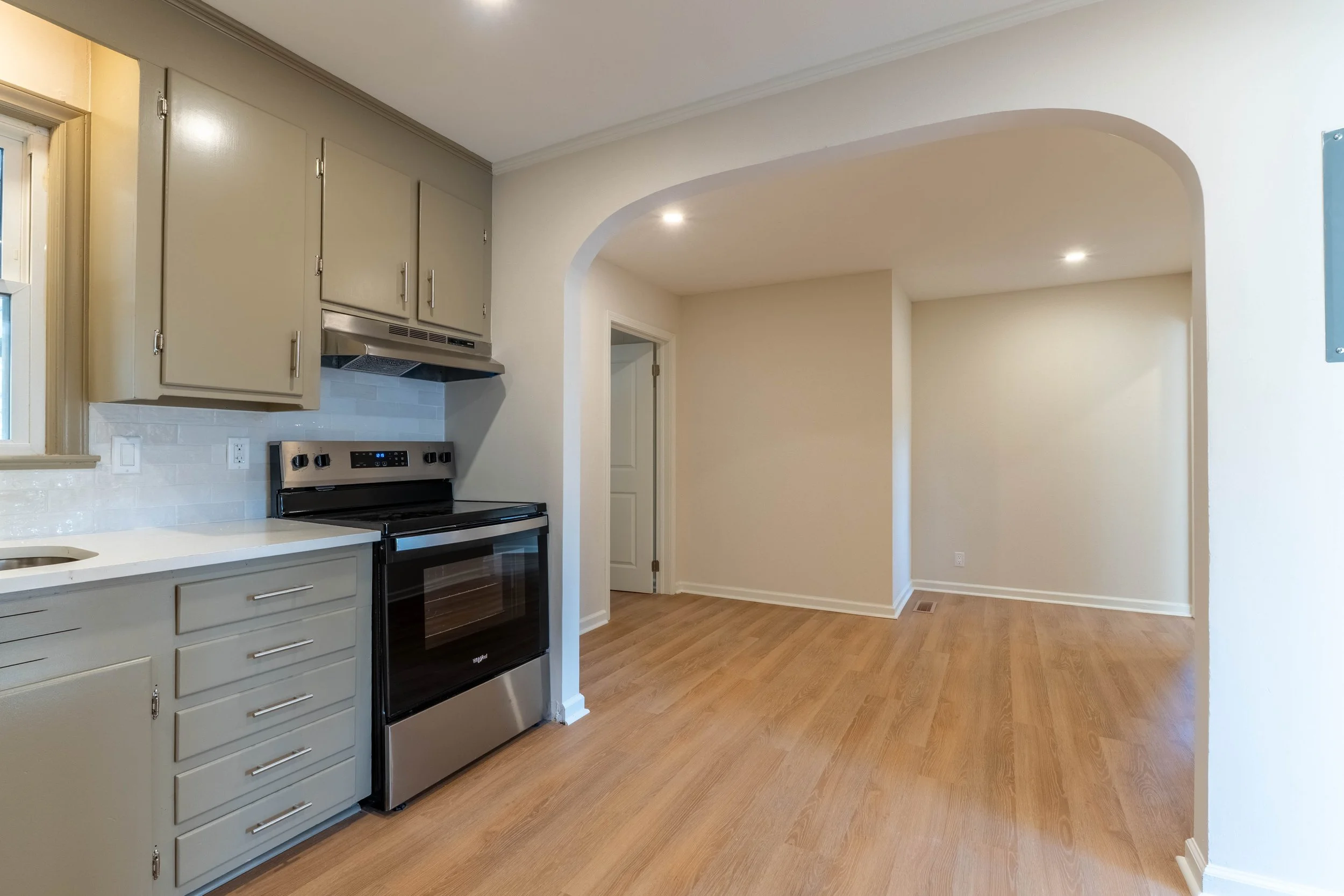 A kitchen with light gray cabinets, a black and stainless steel stove, a white countertop, and wooden flooring. There is an arched doorway leading to an empty room with off-white walls and wooden floors.