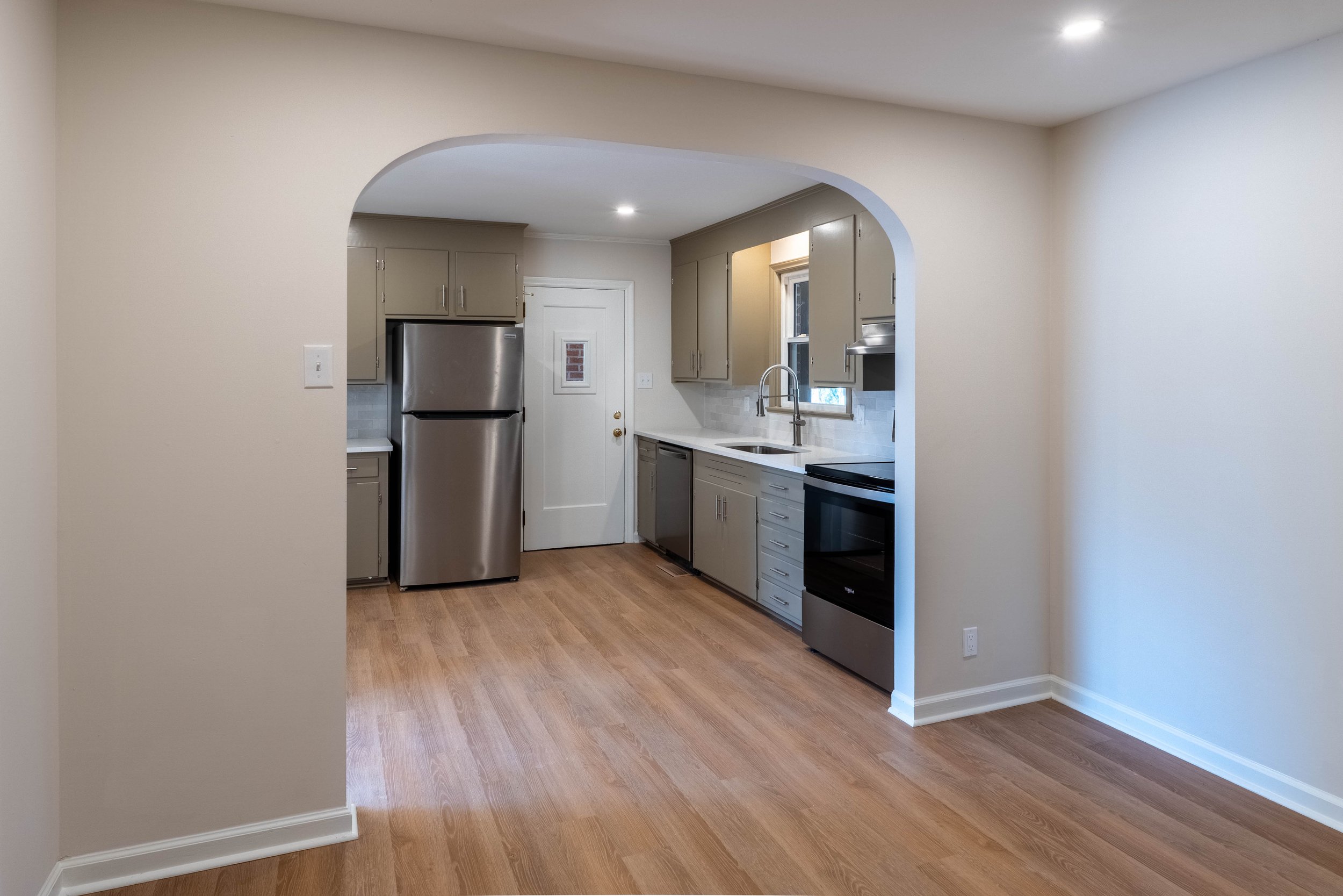 A view of a kitchen with beige cabinets, a stainless steel refrigerator, a white countertop, a sink under a window, and a black oven, through an arched opening in a beige room with wood flooring.