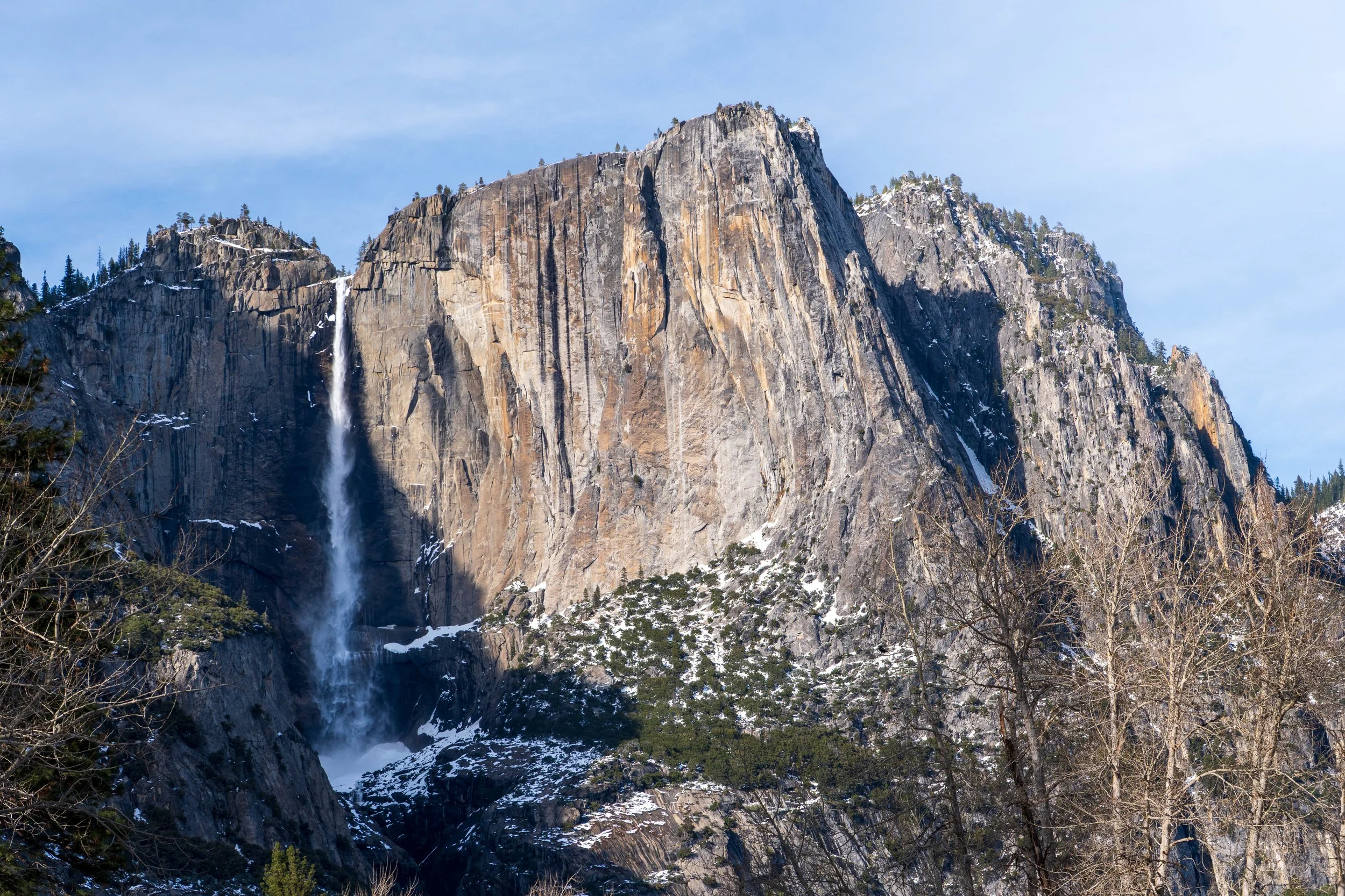 A large granite mountain with a waterfall flowing down its side, with patches of snow and scattered trees at the base and a clear blue sky above.