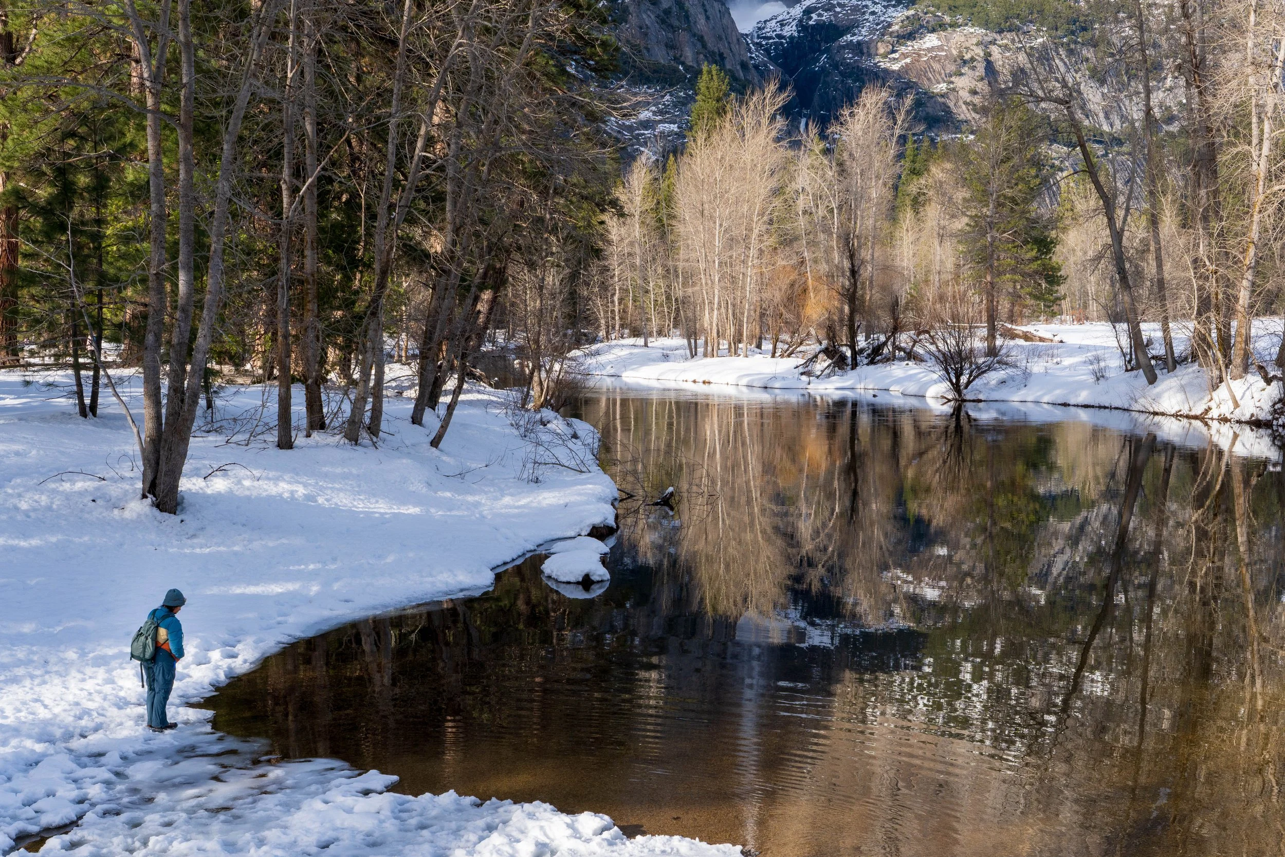A person standing on snow-covered ground near a river in a forested area during winter, with snow-capped mountains in the background.