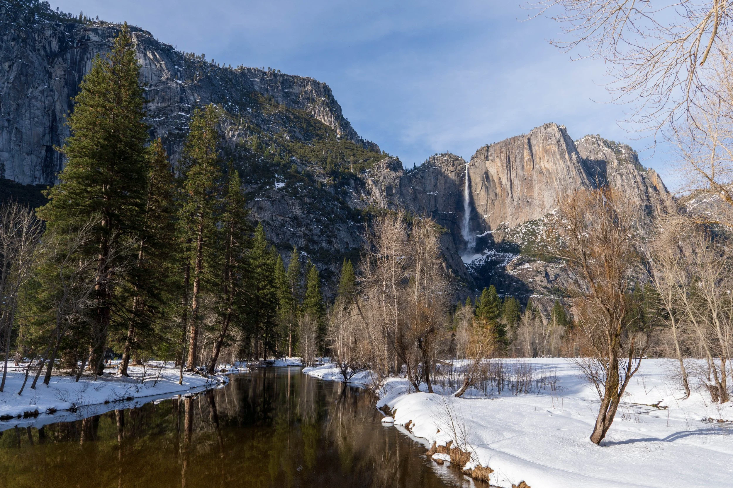 A snowy landscape featuring a calm river in the foreground, with tall trees along the riverbank. In the background, there are large granite cliffs with patches of snow and a thin waterfall cascading down the mountain.