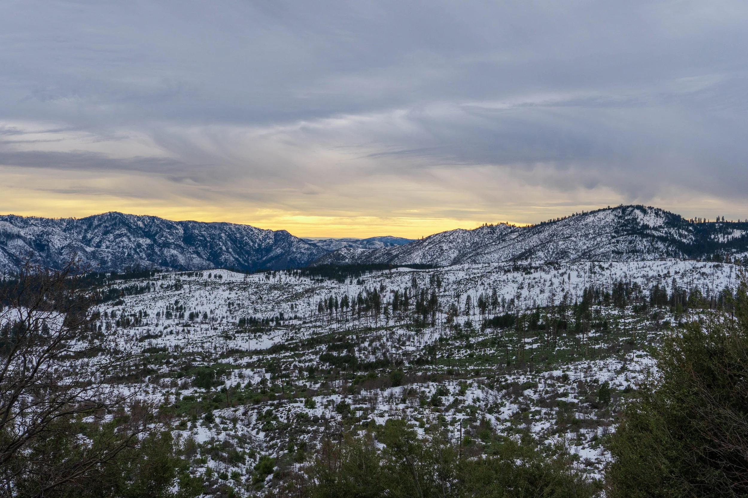 Scenic view of snow-covered mountains under cloudy sky at sunset or sunrise with green trees in the foreground.