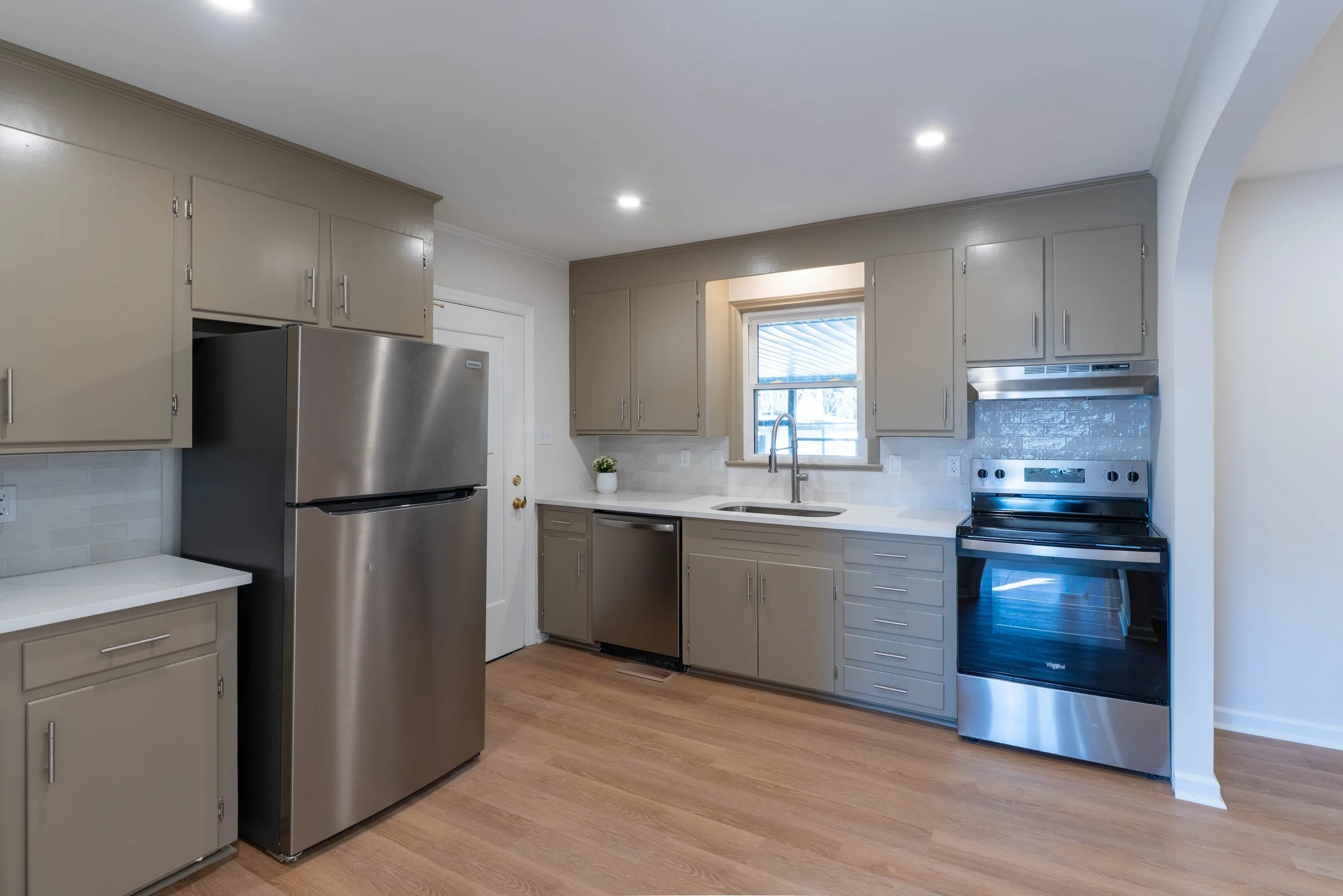 A modern kitchen with beige cabinets, a stainless steel refrigerator, a stainless steel stove, and light wooden flooring. Sunlight comes through a window above the sink, which has a potted plant on the counter nearby.