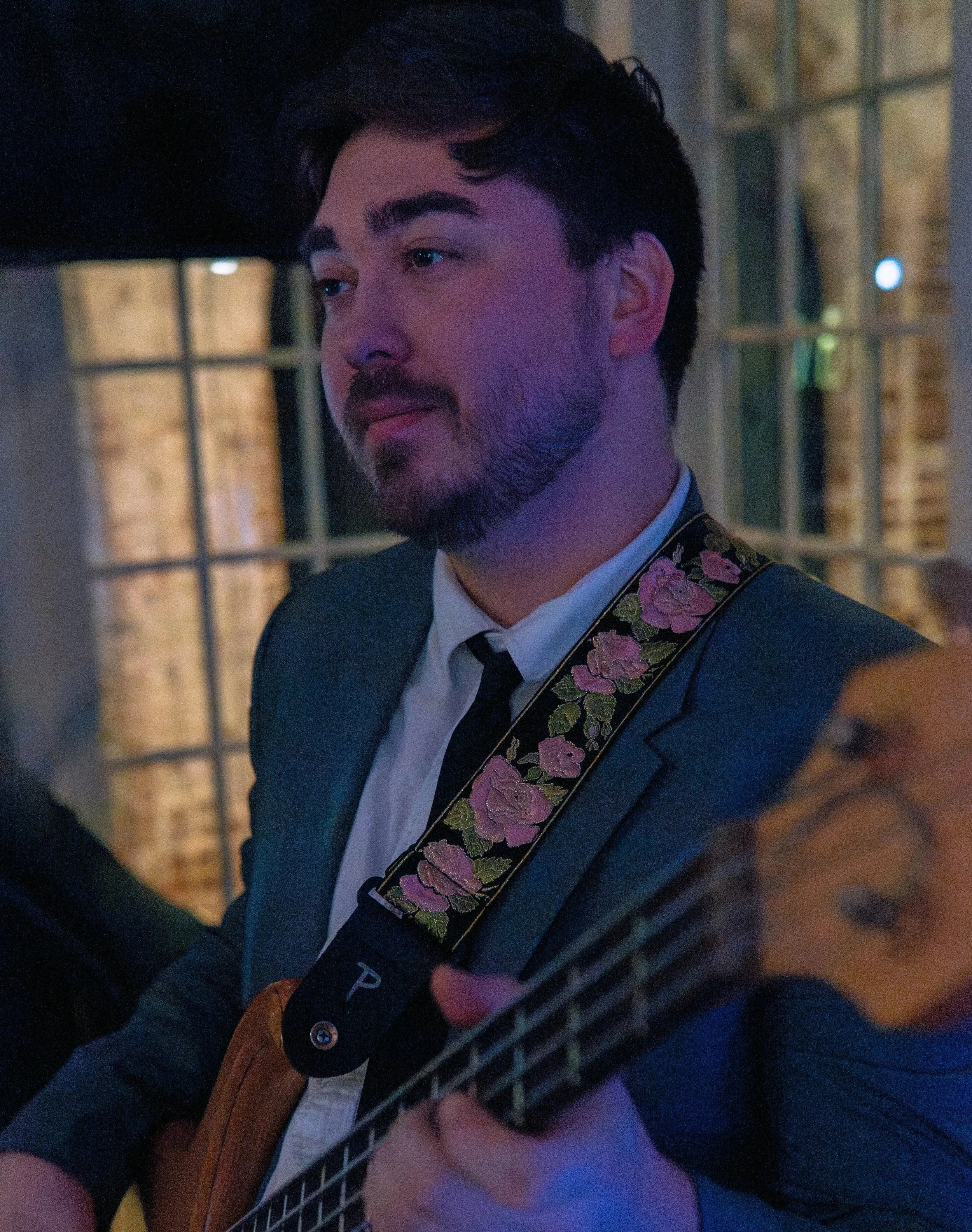 A man with dark hair and a beard playing an electric guitar with a floral strap in a dimly lit room.