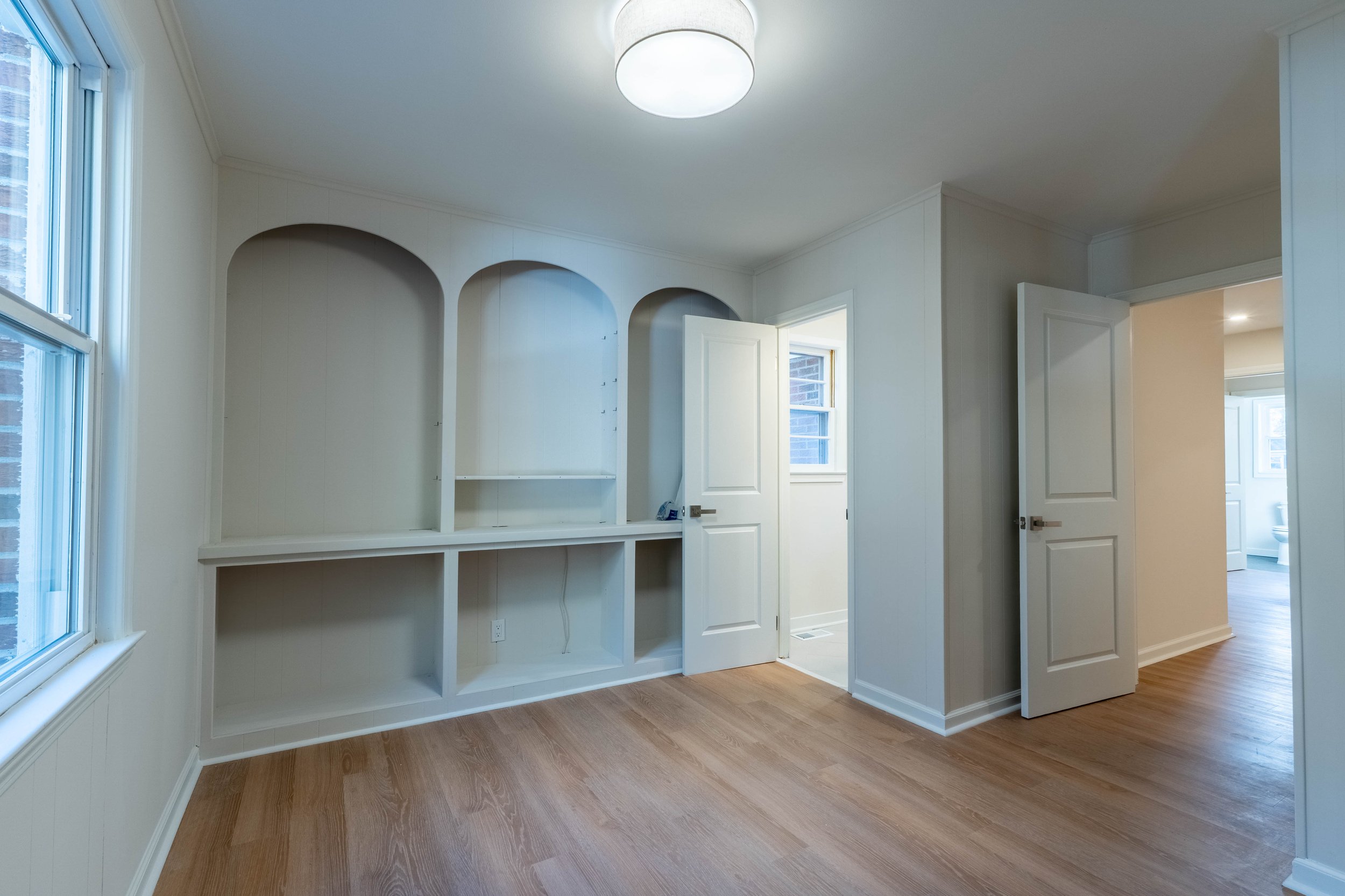 Empty room with beige walls and wood flooring, featuring a built-in white bookshelf and large windows letting in natural light.