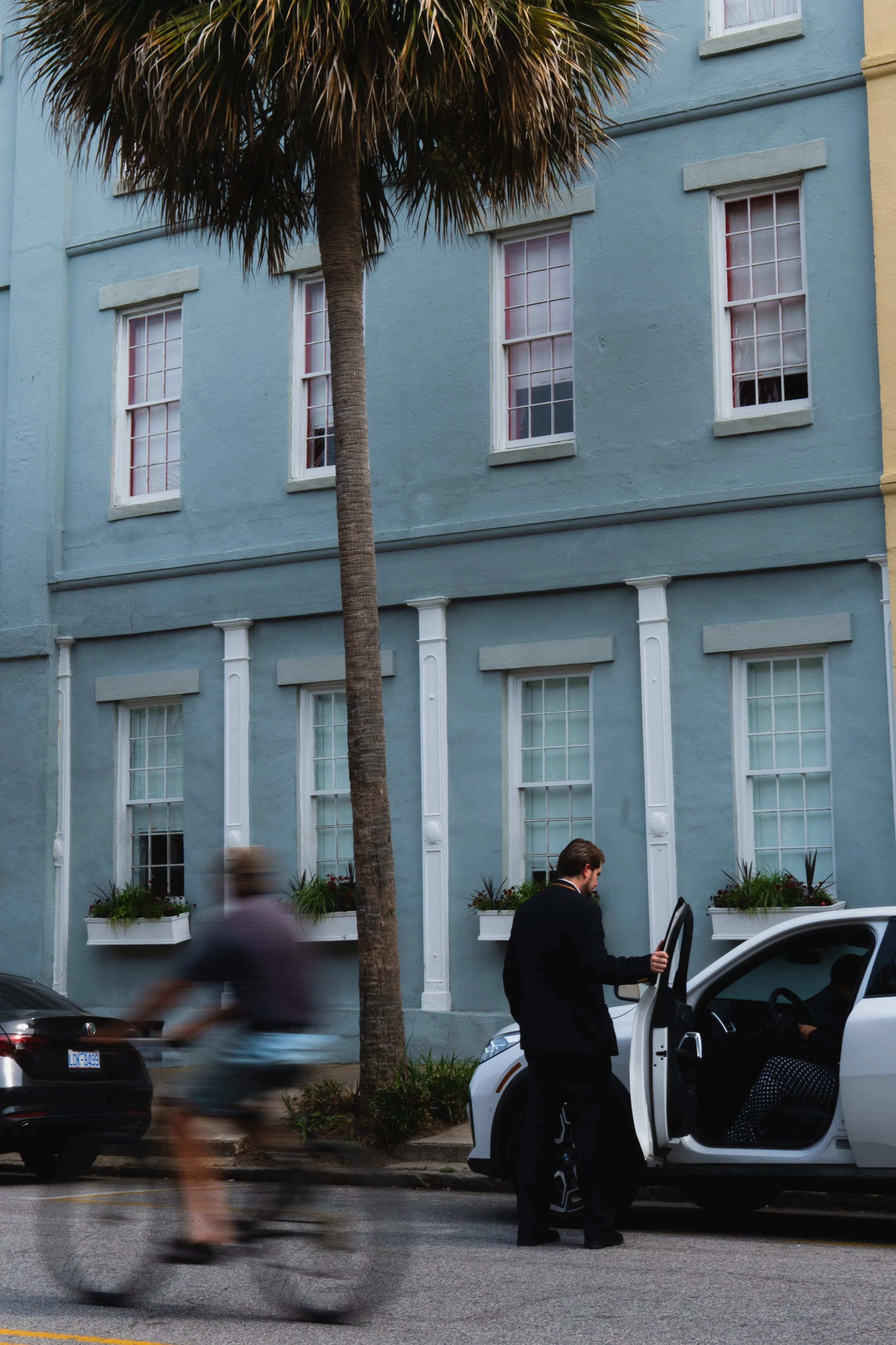 People walking and getting into a white electric vehicle parked on the street in front of a blue building with white window frames and flower boxes, with a palm tree in the foreground.