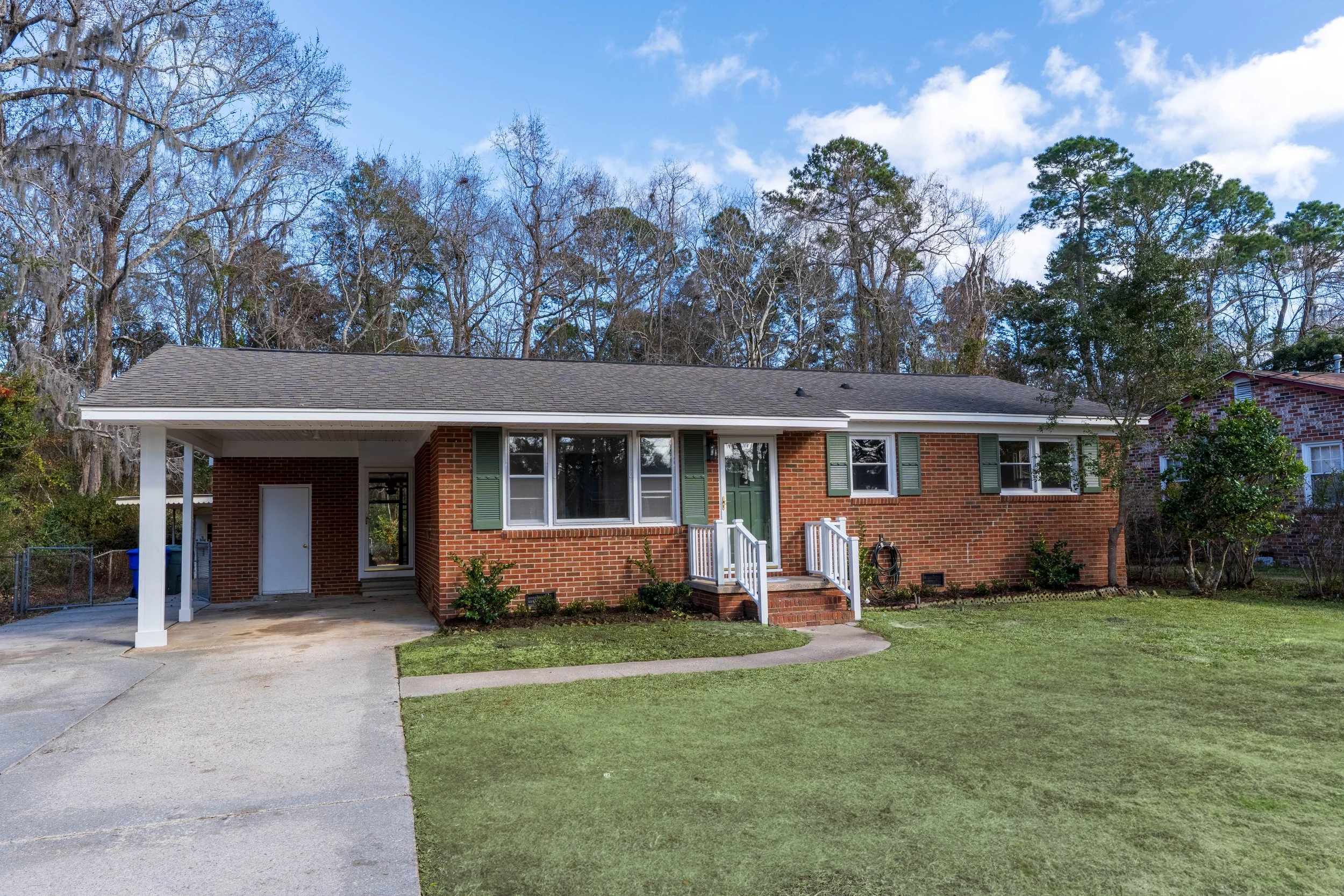 Back of a brick house with a porch, green shutters, small steps, and a driveway, surrounded by a lawn and trees in the background under a partly cloudy sky.