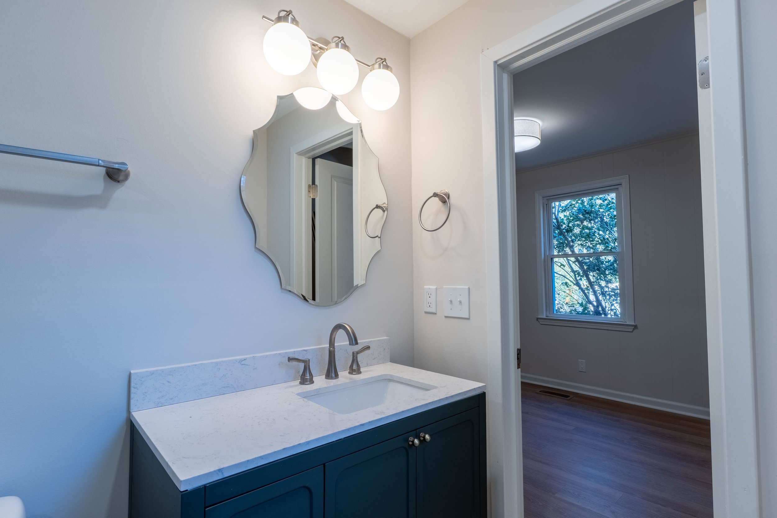 Bathroom with white walls, a vanity with a white countertop, a silver faucet, a decorative mirror, a light fixture with three bulbs, and a window showing trees outside.