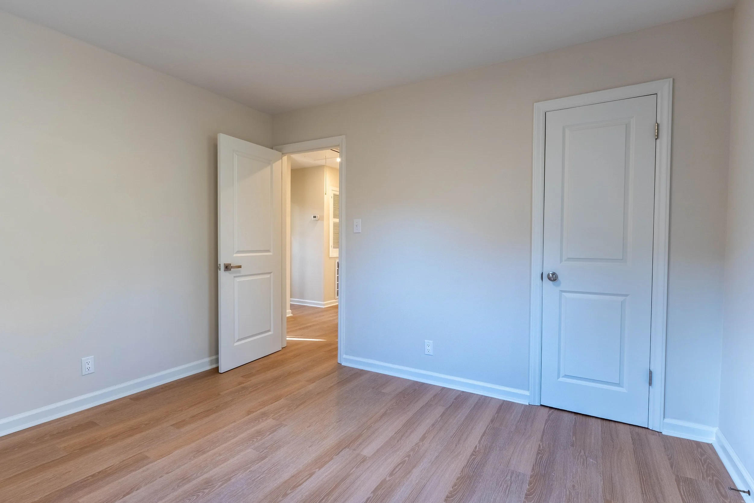 Empty room with white walls, hardwood floors, two white doors, one slightly open revealing a hallway, and visible electrical outlets.
