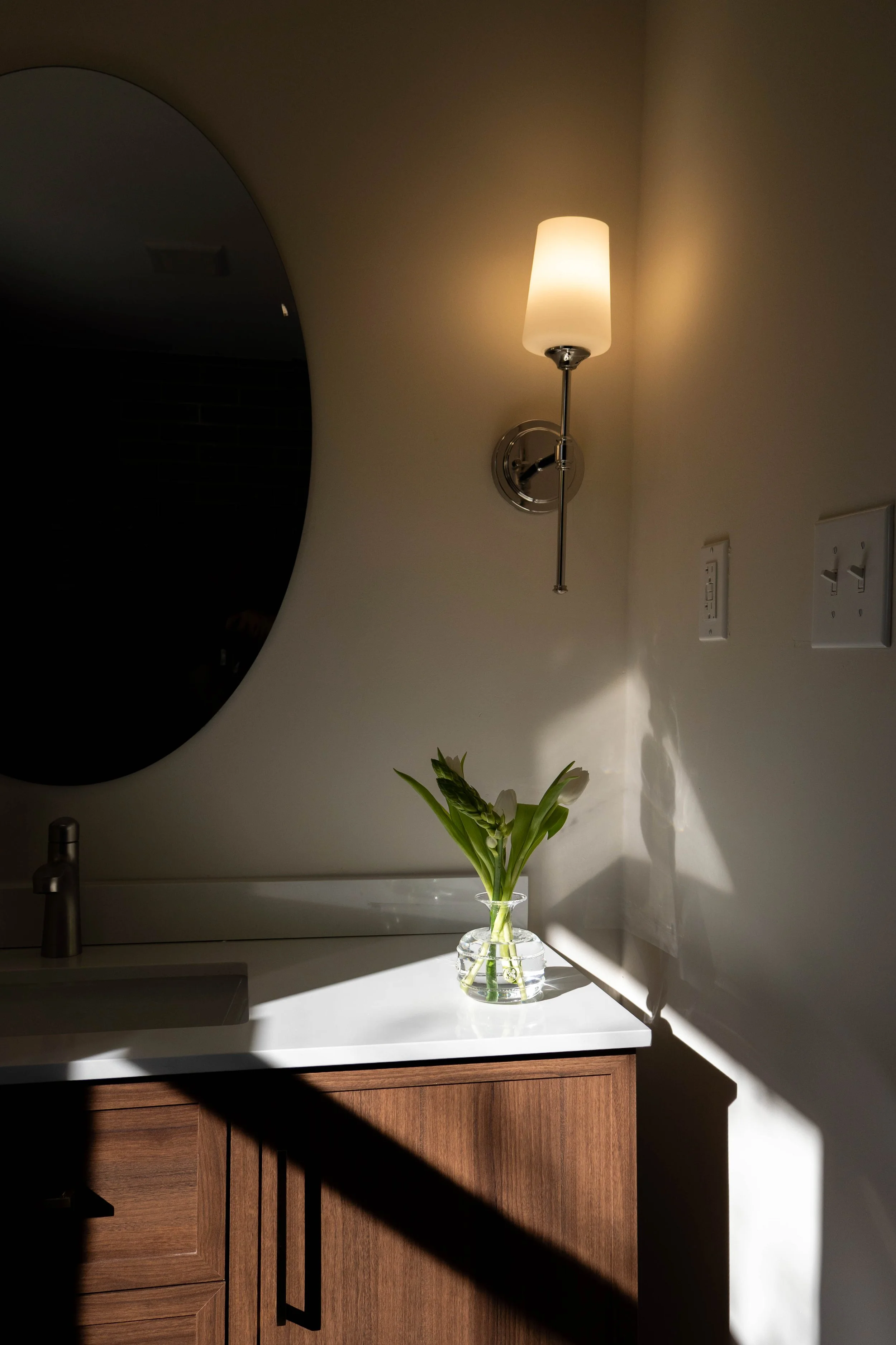 Bathroom vanity with a white countertop, a wooden cabinet, a silver faucet, a flower vase with white flowers, a wall-mounted light fixture turned on, a mirror, and an electrical outlet on the wall.