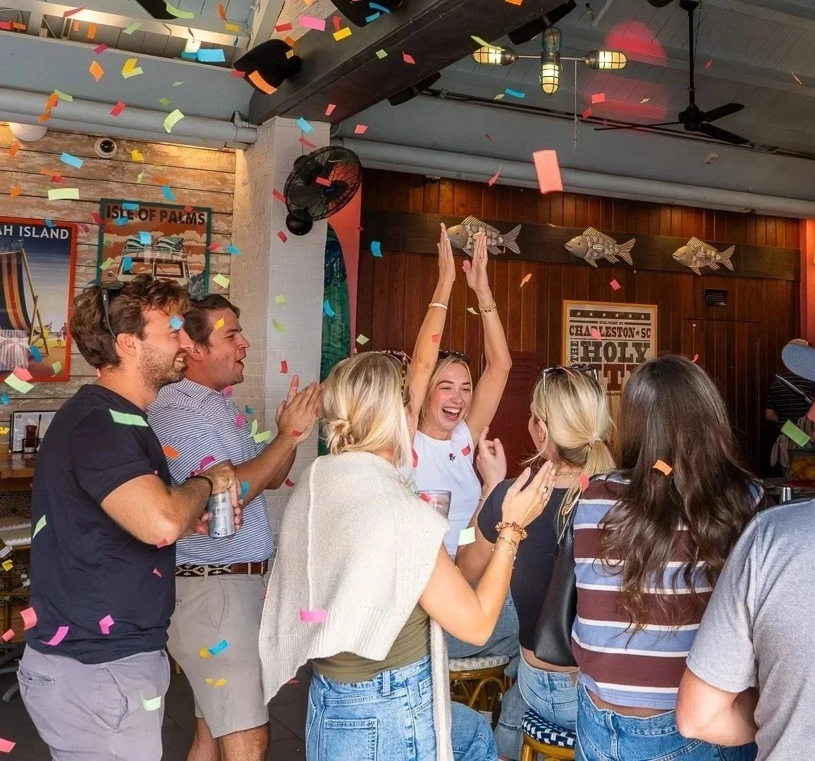 A group of people celebrating indoors with confetti falling, some with their hands raised, smiling and talking to each other.
