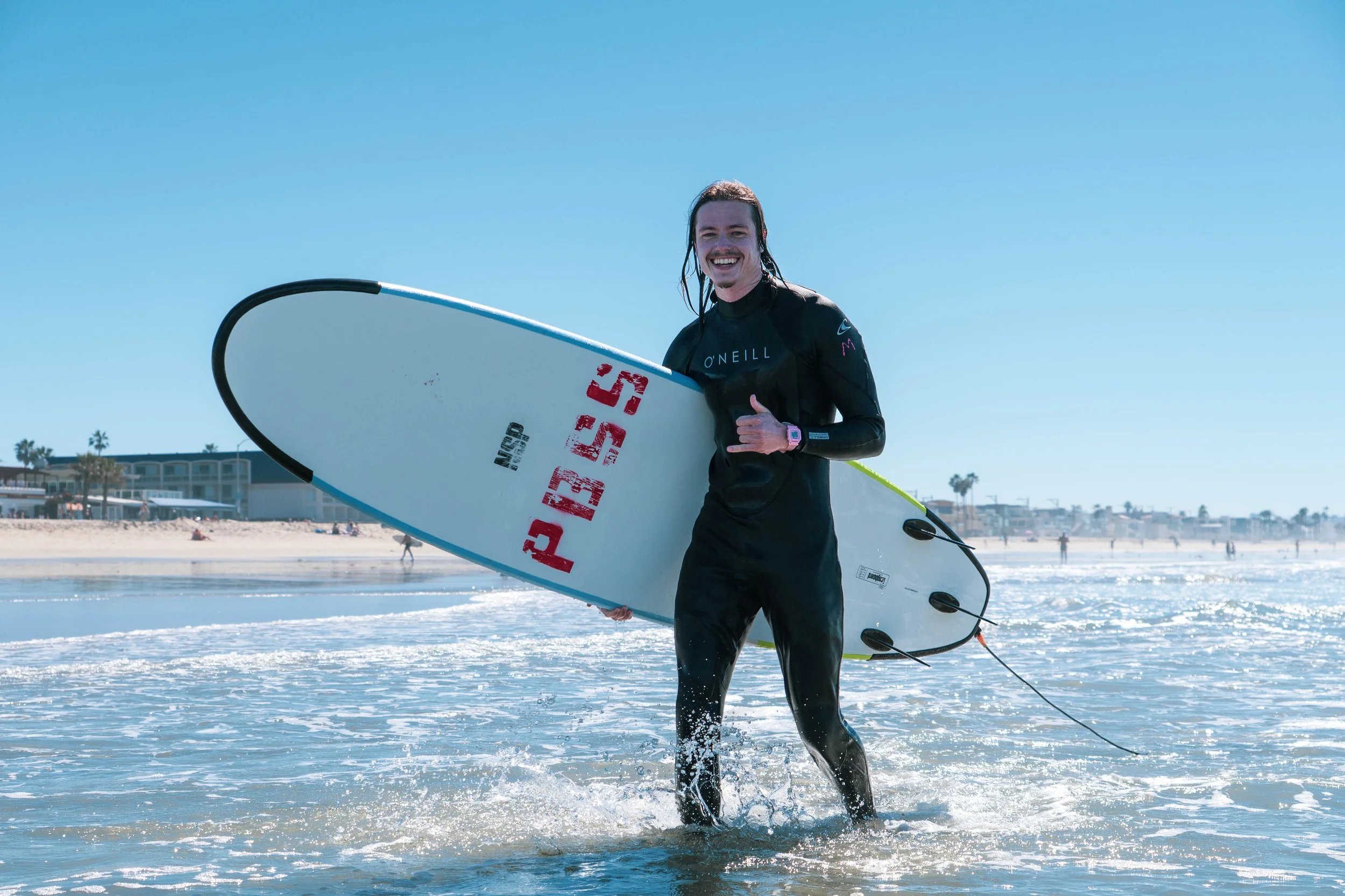A man holding a surfboard while standing in shallow water at the beach, smiling and giving a thumbs-up gesture under a clear blue sky.