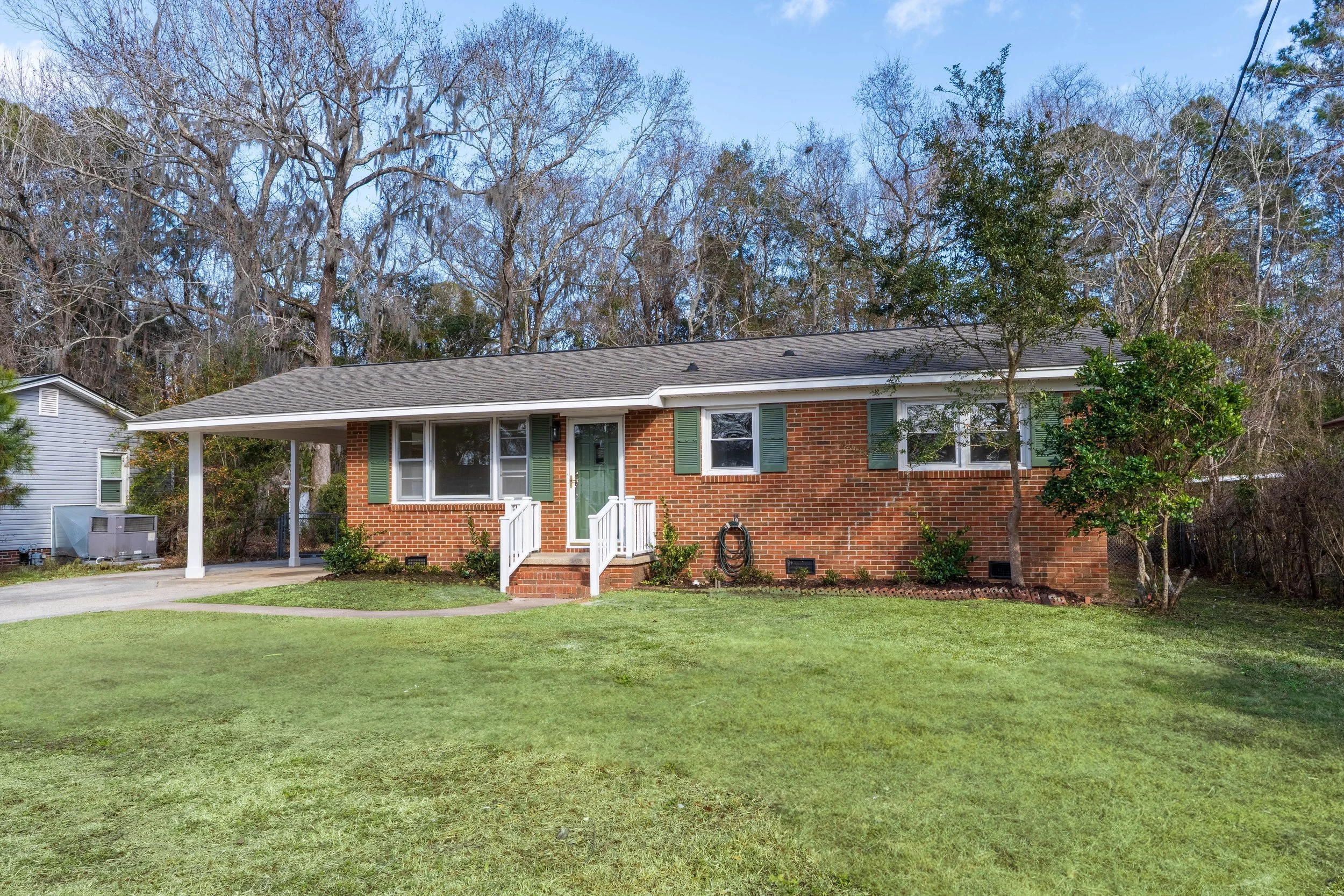 A single-story brick house with a gray roof, green shutters, and white porch stairs, situated on a grassy lawn with trees and a wooded background.