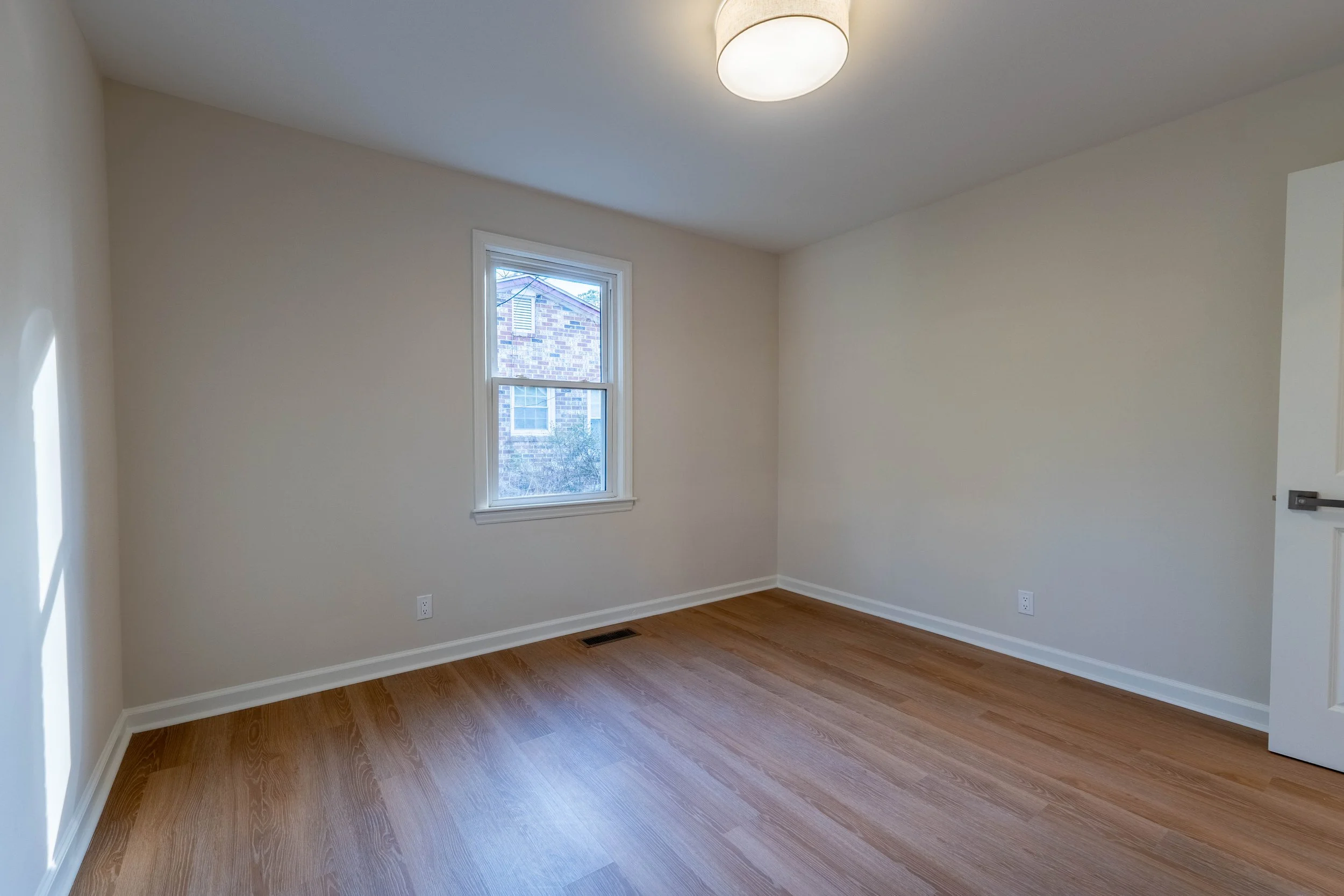 Empty room with beige walls, a window, hardwood floor, and a ceiling light fixture.