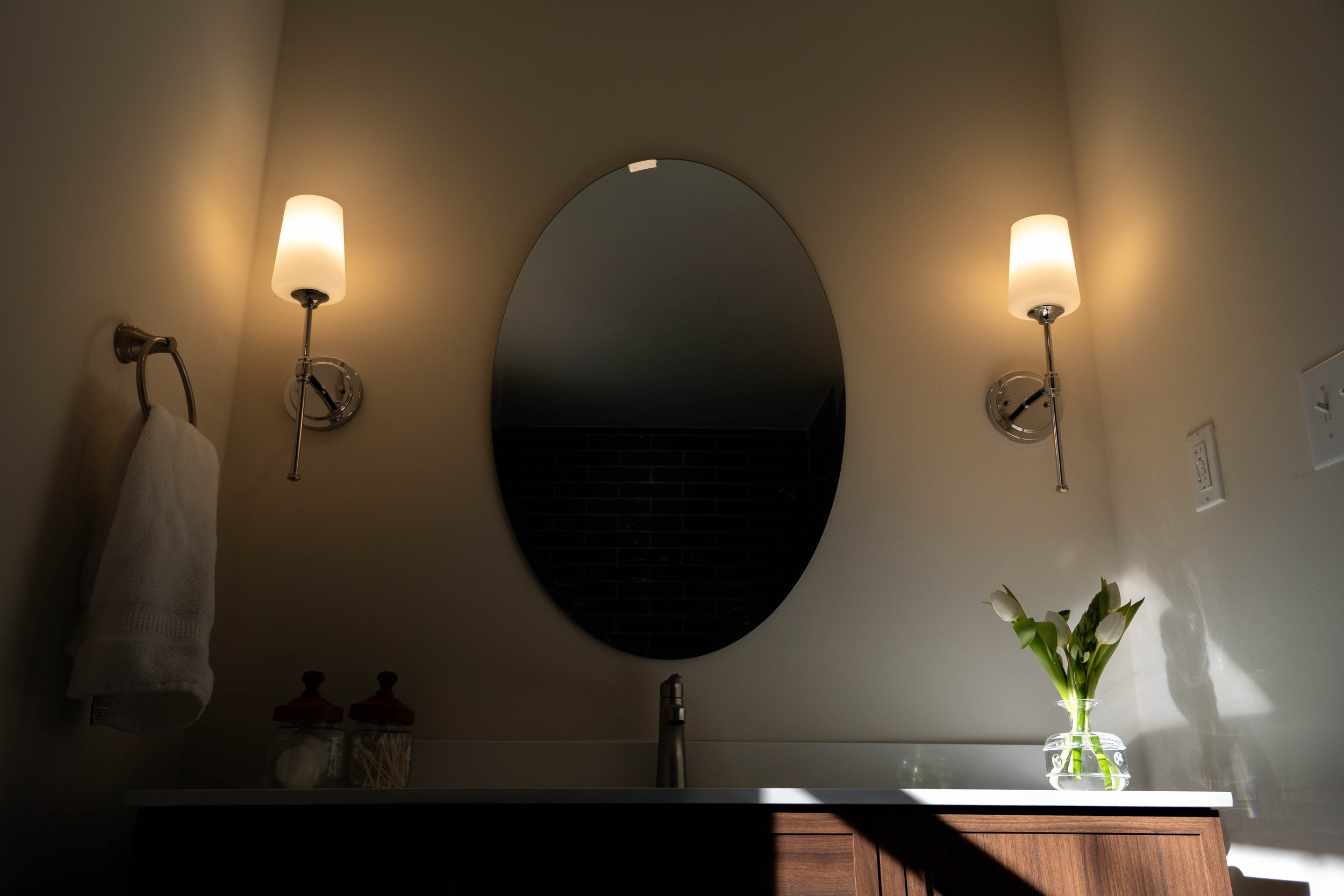 A bathroom vanity with two wall-mounted lamps, an oval mirror, a towel hanging on a wall, a vase with white flowers, and various toiletries on the counter.