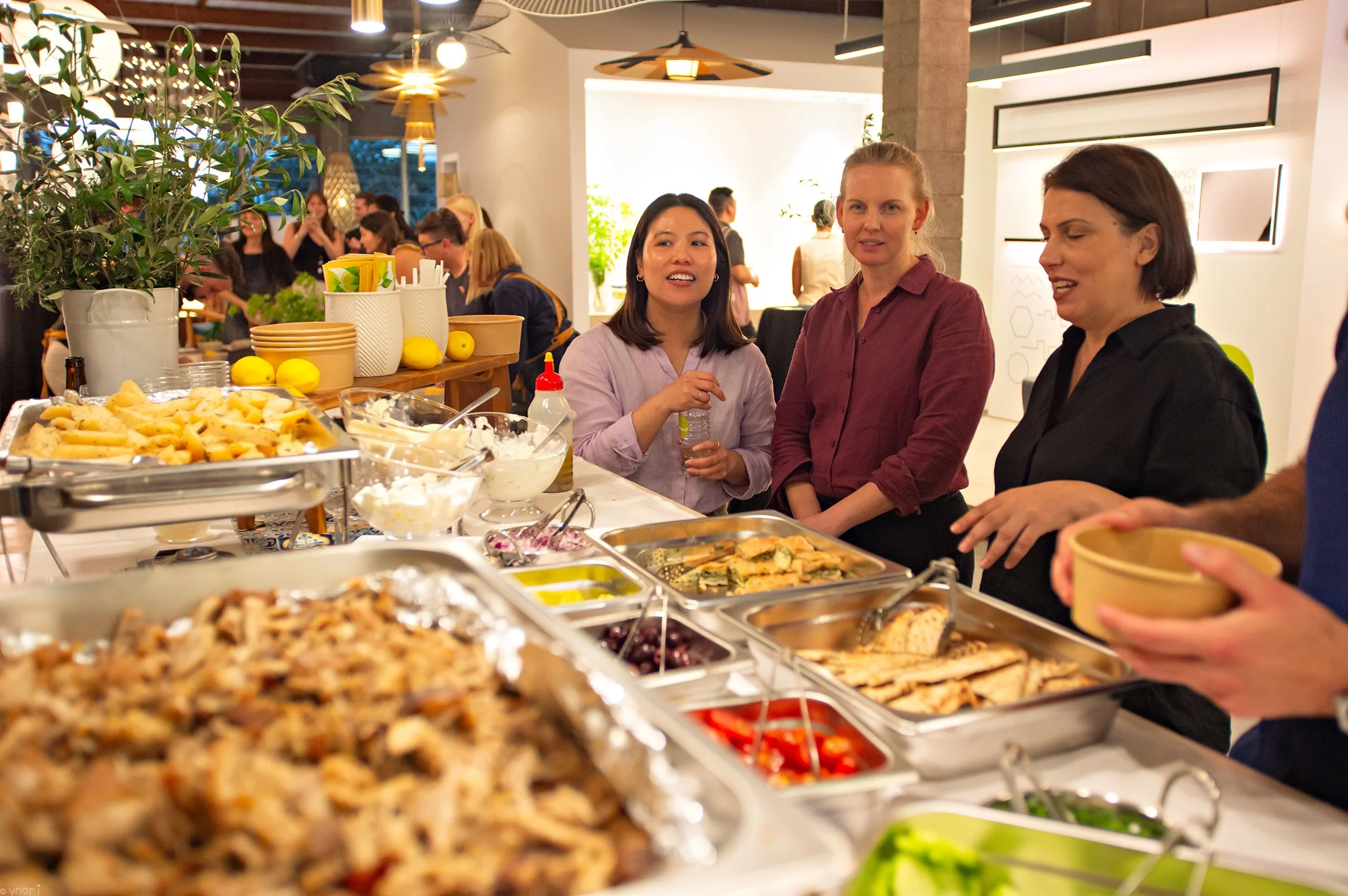 People enjoying a buffet at a social gathering, with various dishes and food items on the table including salads, grilled meats, and sides, in a well-lit, modern indoor setting.