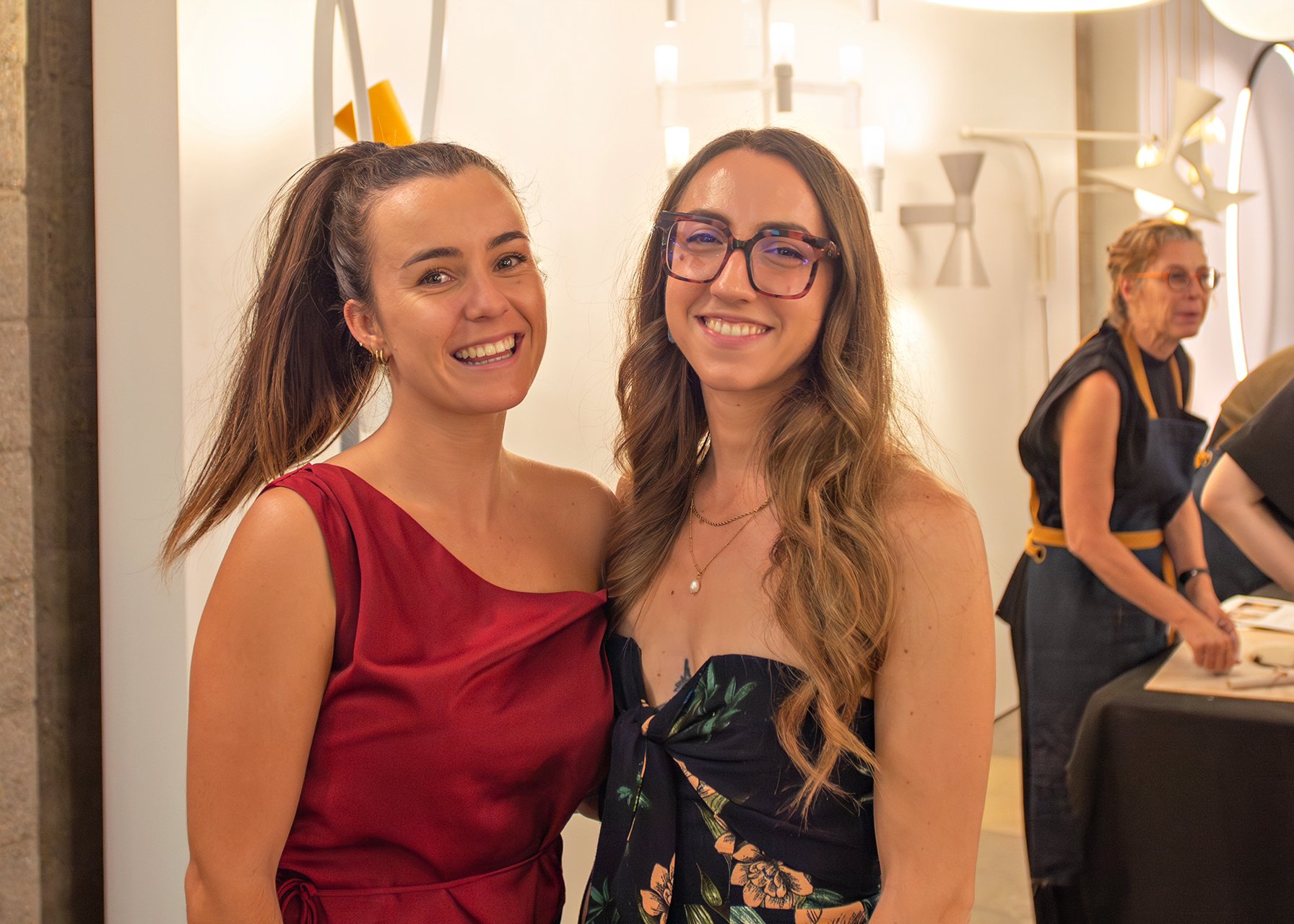 Two smiling women standing close together indoors, with a third woman in the background, at what appears to be a social gathering or event.