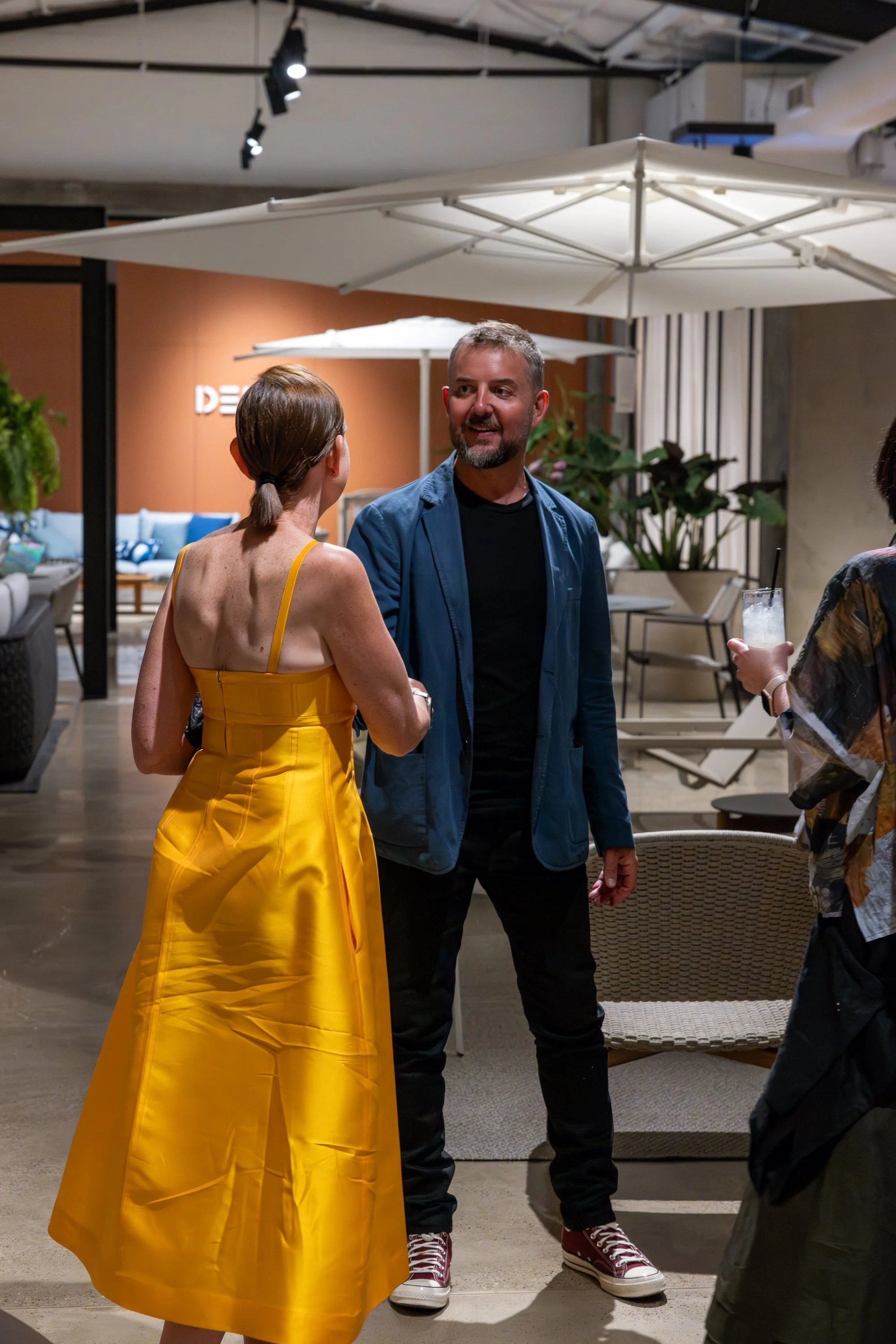 People talking at a social event indoors, with a woman in a bright yellow dress on the left and a man in a blue jacket and black pants in the center. Another person holding a drink is partially visible on the right. There are umbrellas and furniture 